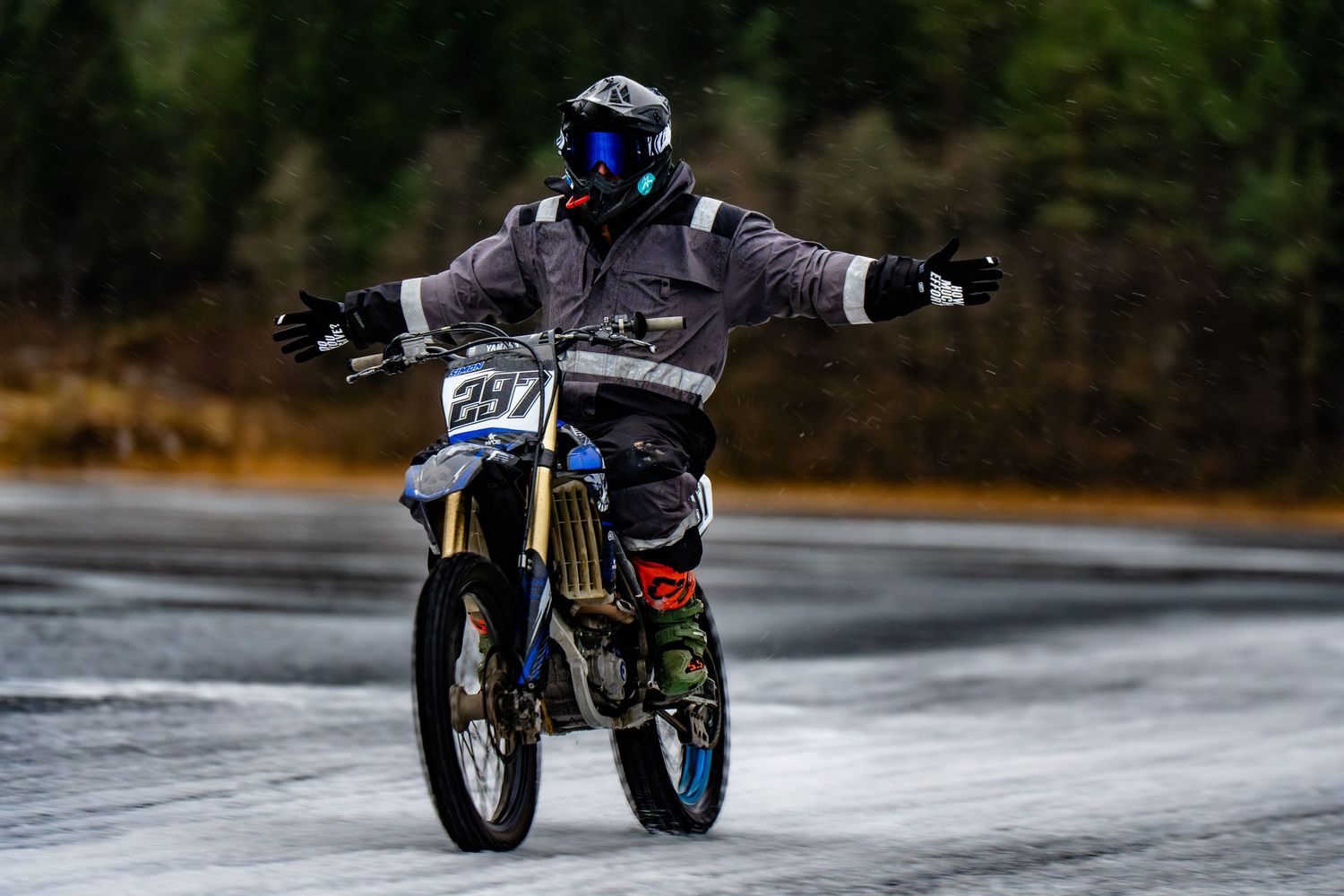 A blue motorcycle performs a dynamic drift maneuver on a snowy road surface, creating a dramatic spray of snow.