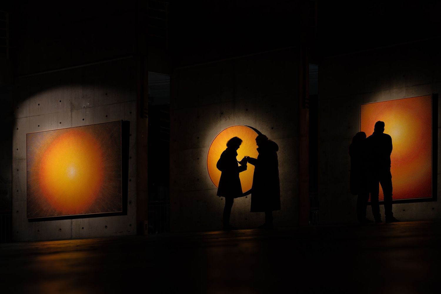 Silhouettes of visitors stand against backlit orange circular artworks in an exhibition.