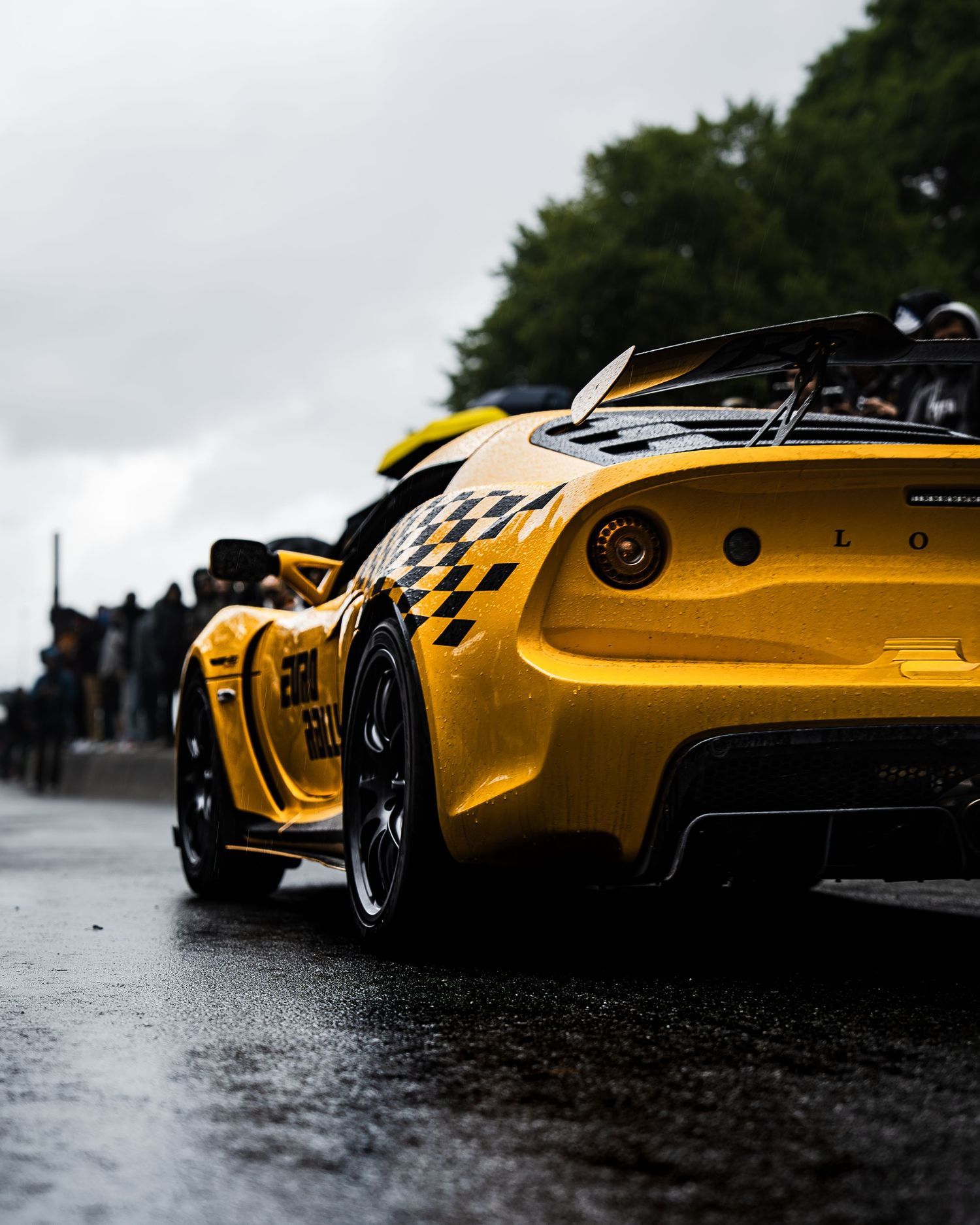 Rear detail shot of yellow sports car with racing checkers on wet track during overcast weather.