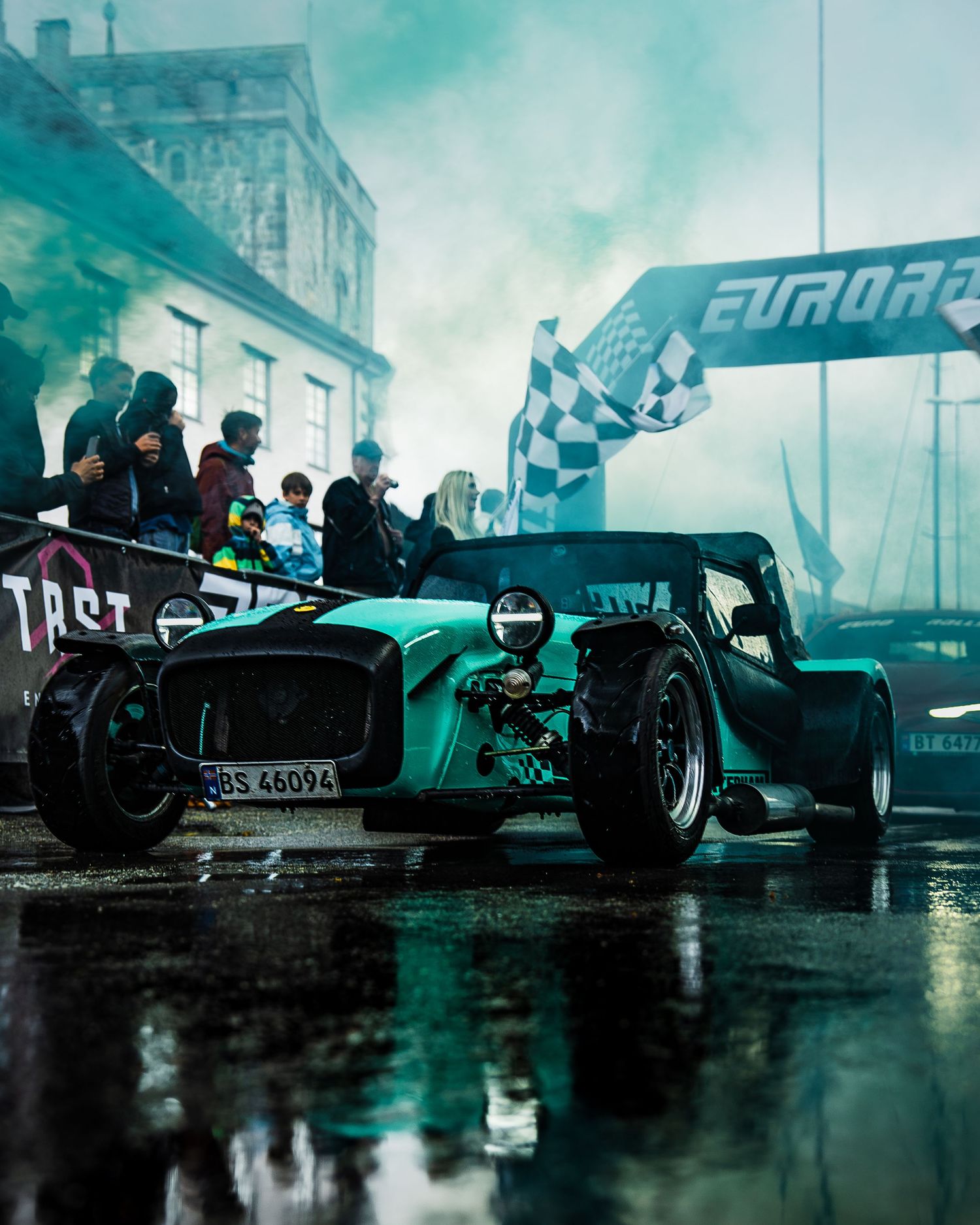 Teal-lit vintage Caterham race car surrounded by spectators at rainy EuroRally event.