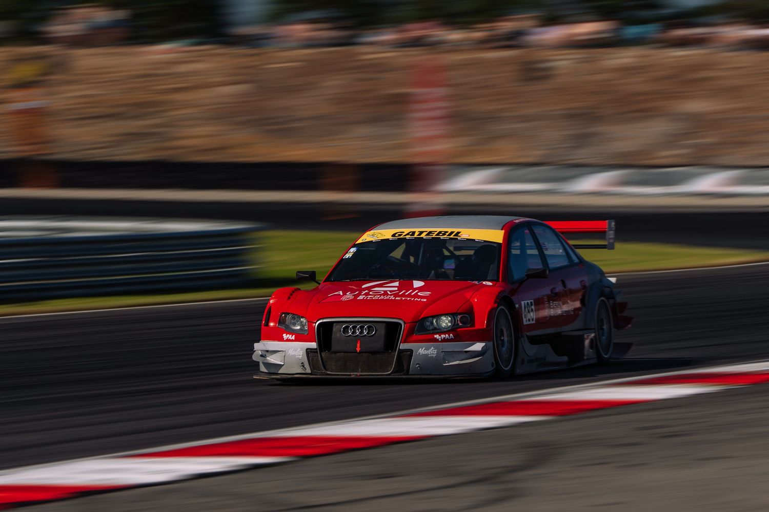 Red race car speeds around track corner with motion blur during competitive motorsport event.