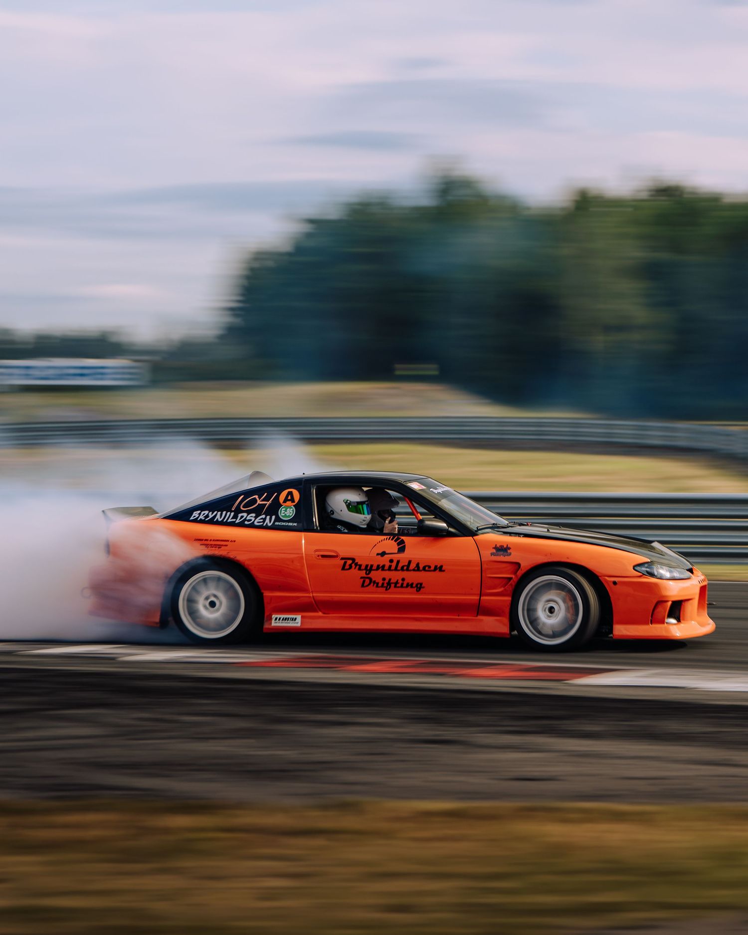 Orange drift car performs a smoky slide on a race track with trees and cloudy sky in the background.