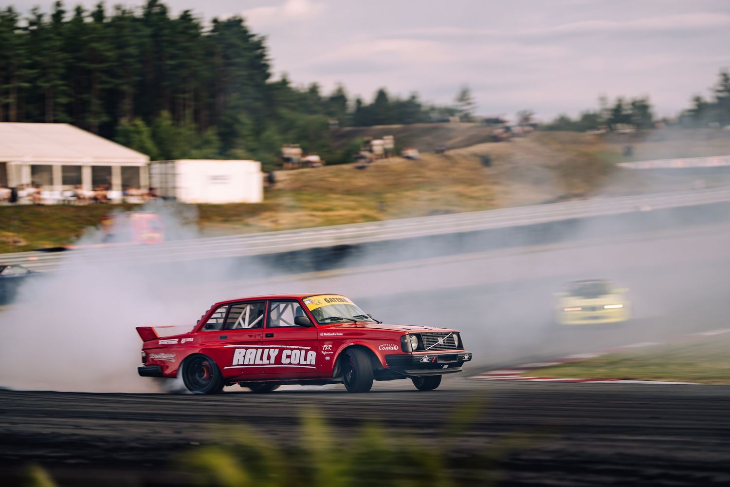 Red vintage car drifting on a racetrack creating smoke clouds against a forest backdrop.