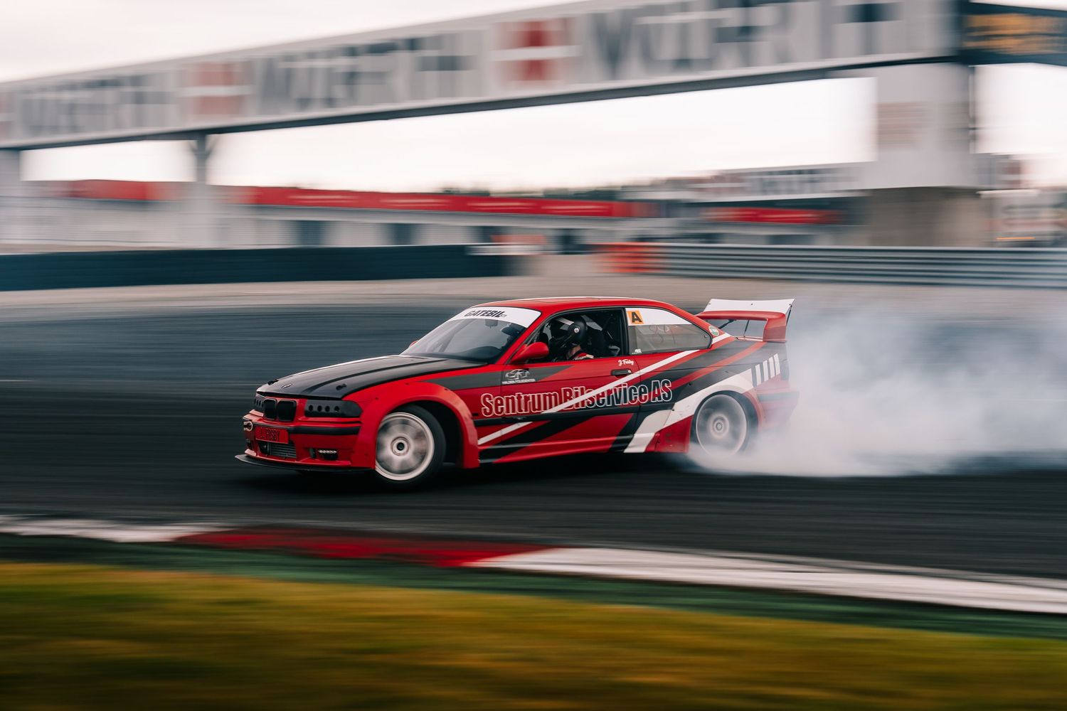 Red sports car drifting on racetrack creating white smoke while performing high-speed maneuvers.