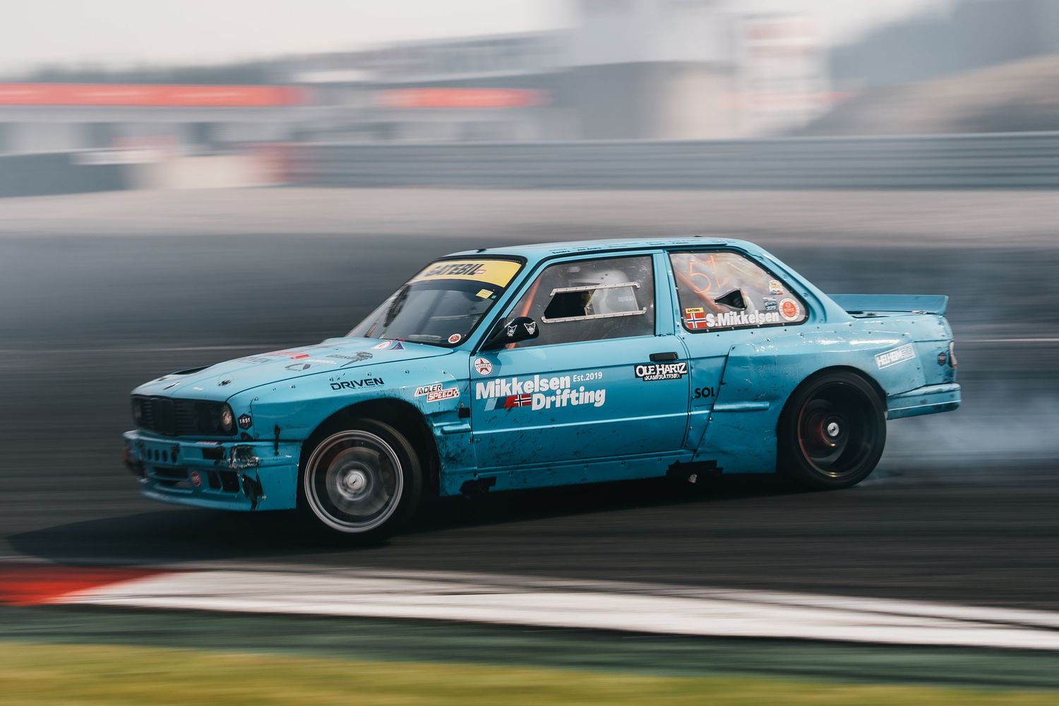 A blue vintage car drifts sideways on a racetrack creating smoke from its tires during a motorsport event.
