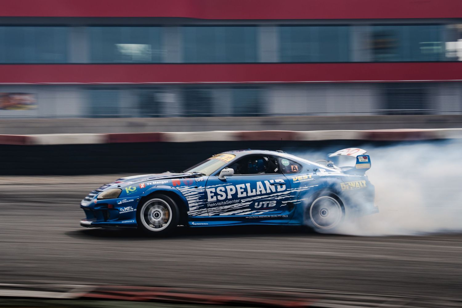 A blue drift car creates smoke while drifting sideways on a racetrack during a motorsport competition.