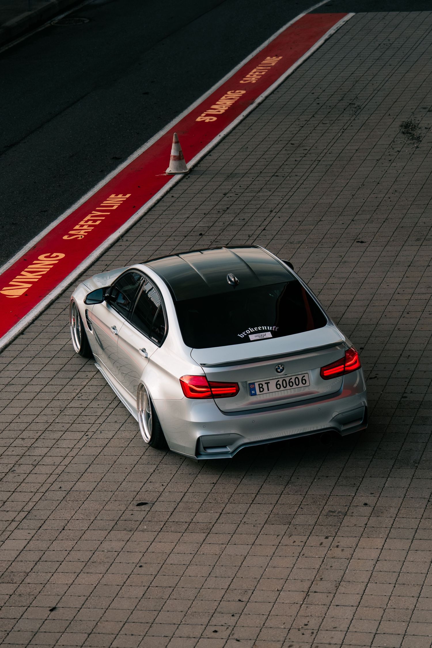 Aerial view of a silver BMW M3 sedan with custom modifications parked on brick pavement next to red racing stripes.