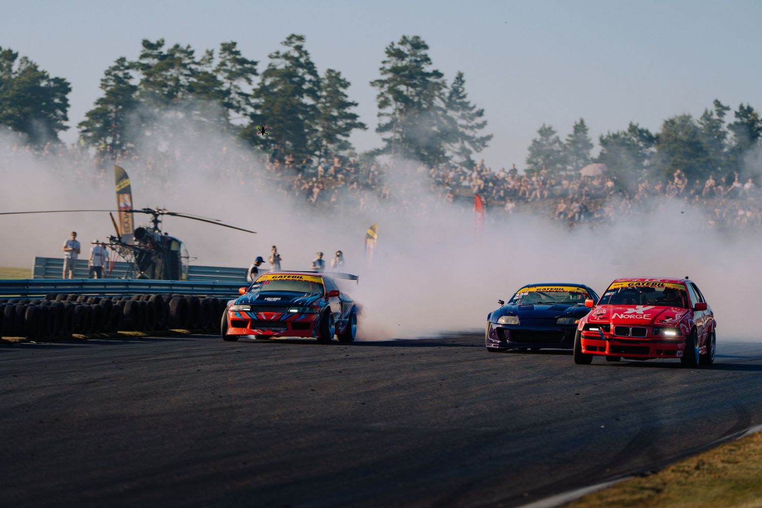 Three drift cars compete on a racetrack creating clouds of tire smoke during an intense drifting competition.
