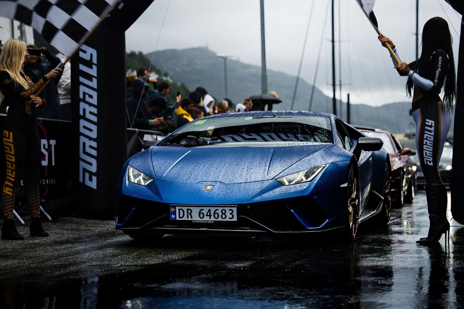 Blue Lamborghini supercar in the rain at a racing event with checkered flags.