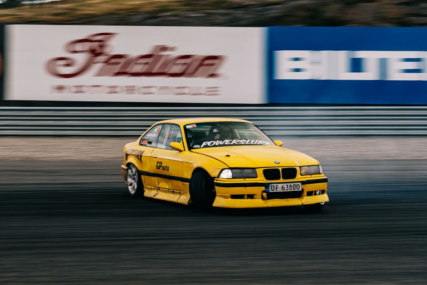 Yellow BMW race car drifting on a race track with smoke trailing from the tires during competition.