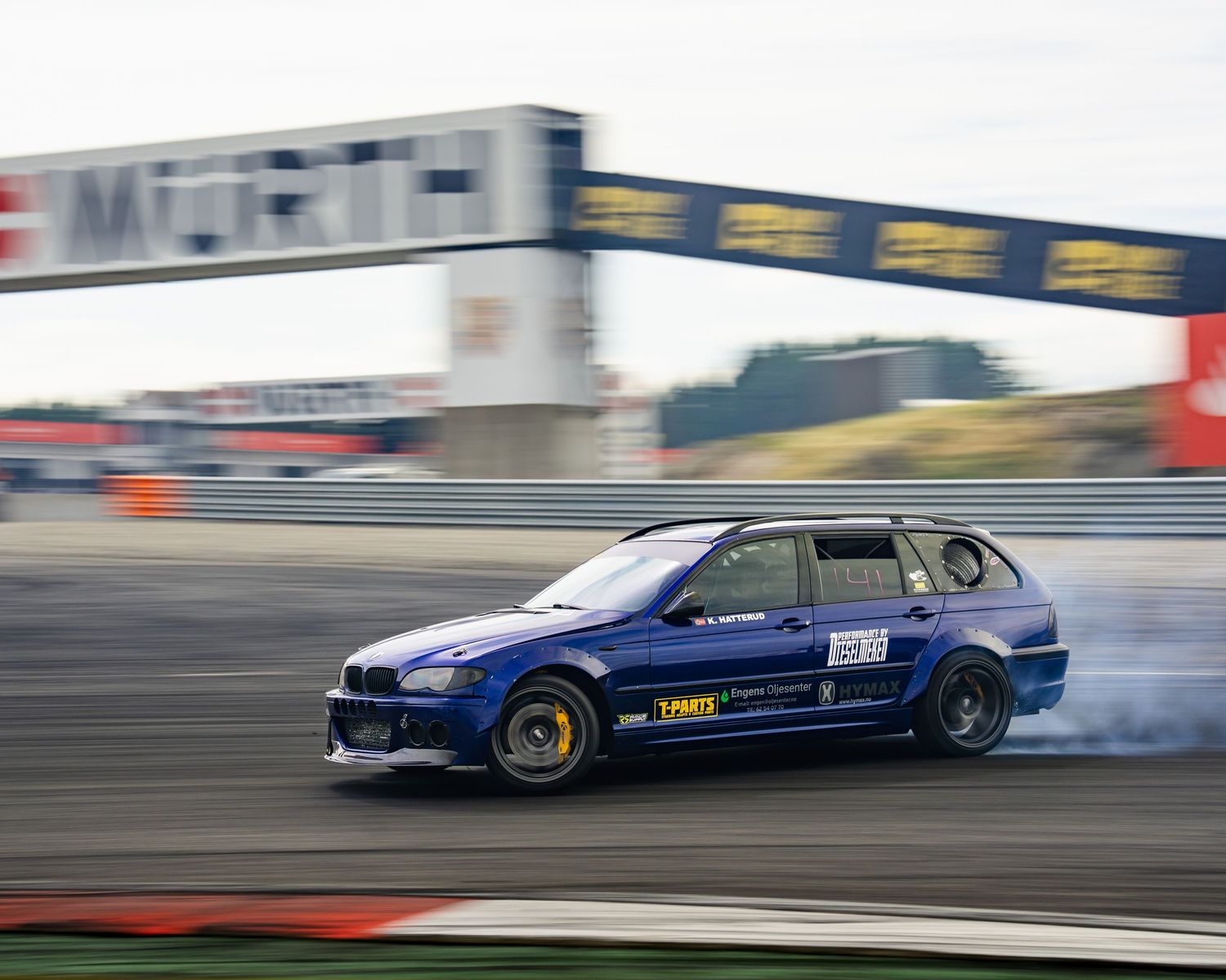 A blue sports car drifts sideways on a race track with smoke coming from the tires during a drifting competition.