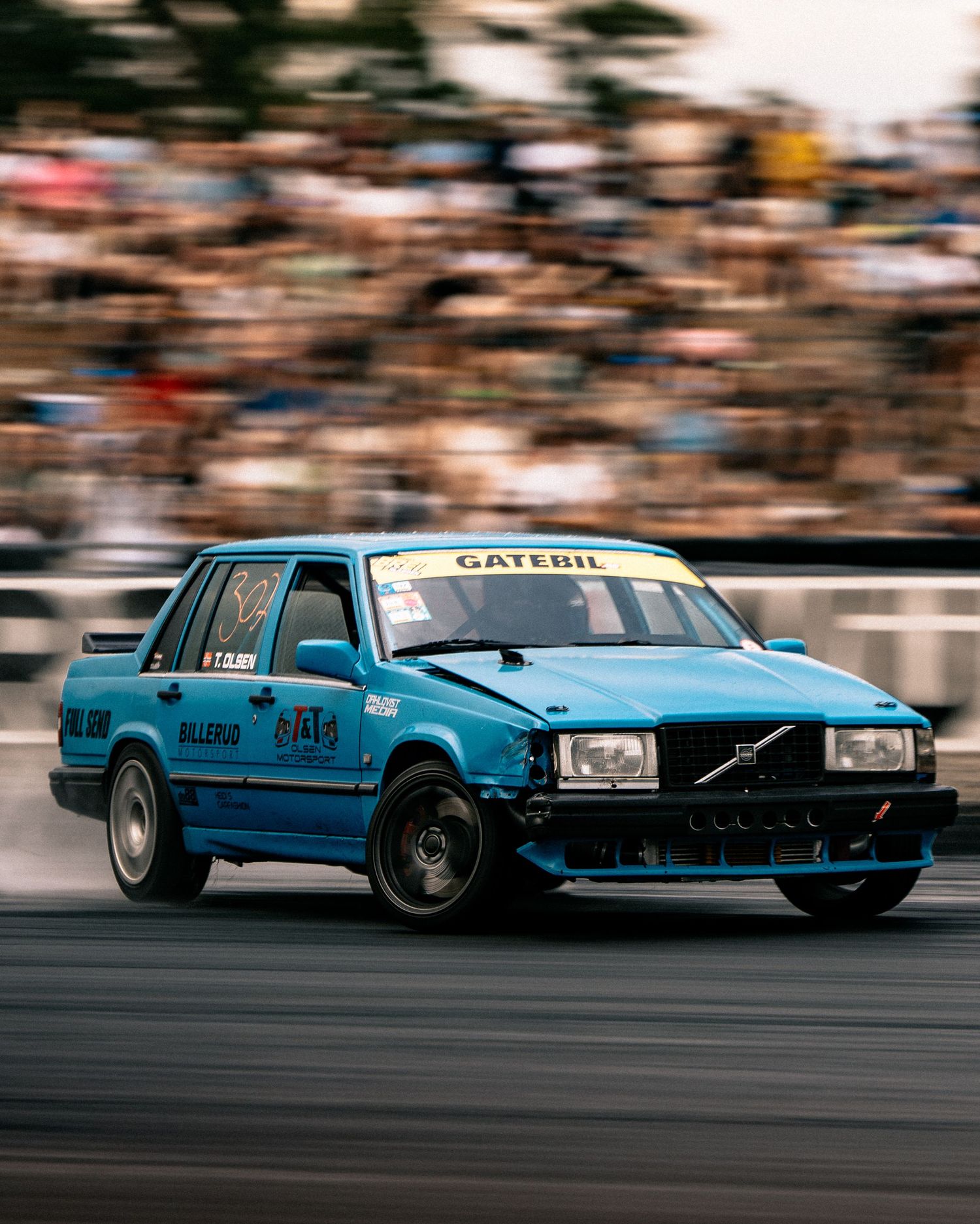 A vintage blue Volvo racing car speeds along a racetrack with motion blur and spectators in the background.