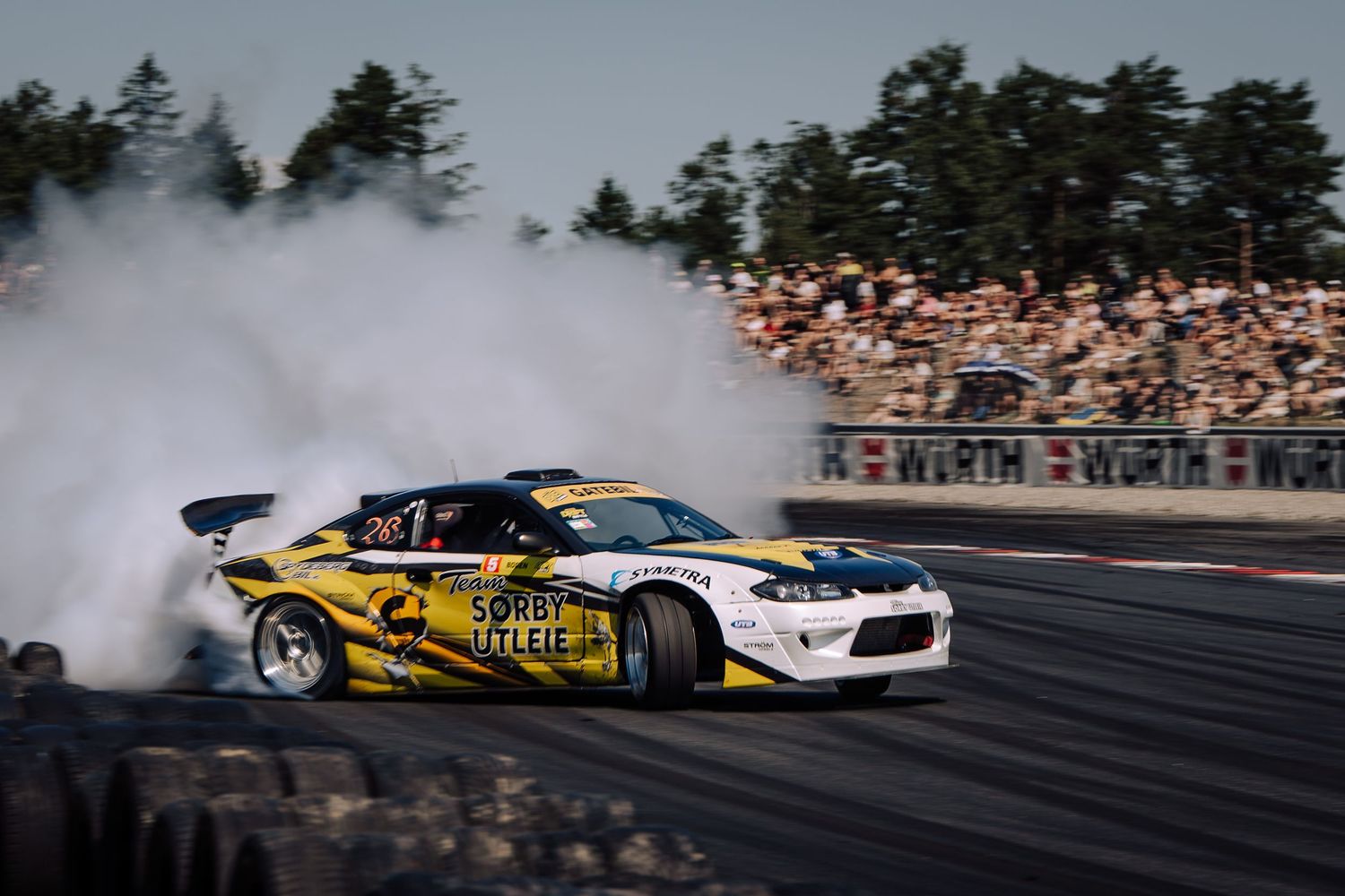 A series of drift cars sliding sideways on a racetrack with smoke billowing from their tires during a competition event.