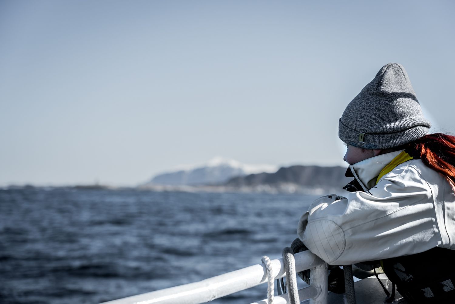 Person in winter gear looking out over the ocean from a boat with mountains in the distance.