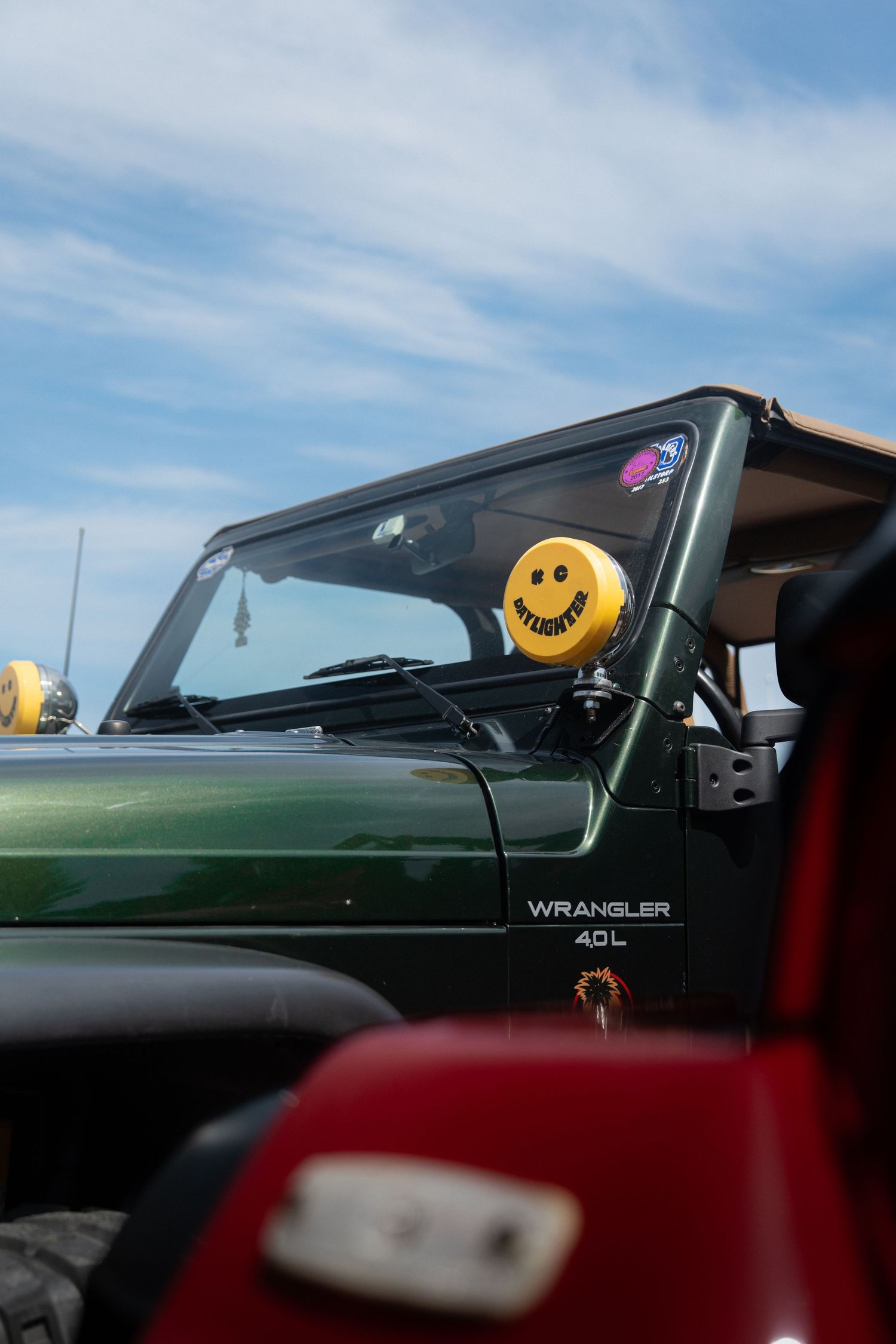 Close up view of a green Jeep Wrangler with yellow smiley face antenna toppers against blue sky.