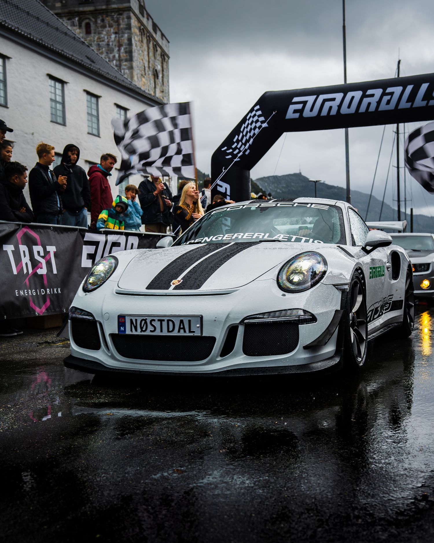 White Porsche 911 GT3 RS with racing stripes under Eurorally finish arch in wet conditions.