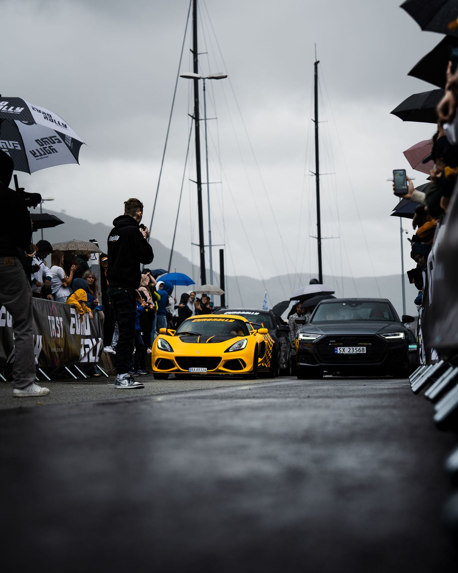 Yellow sports car driving through crowd of spectators with umbrellas at a rainy racing event.