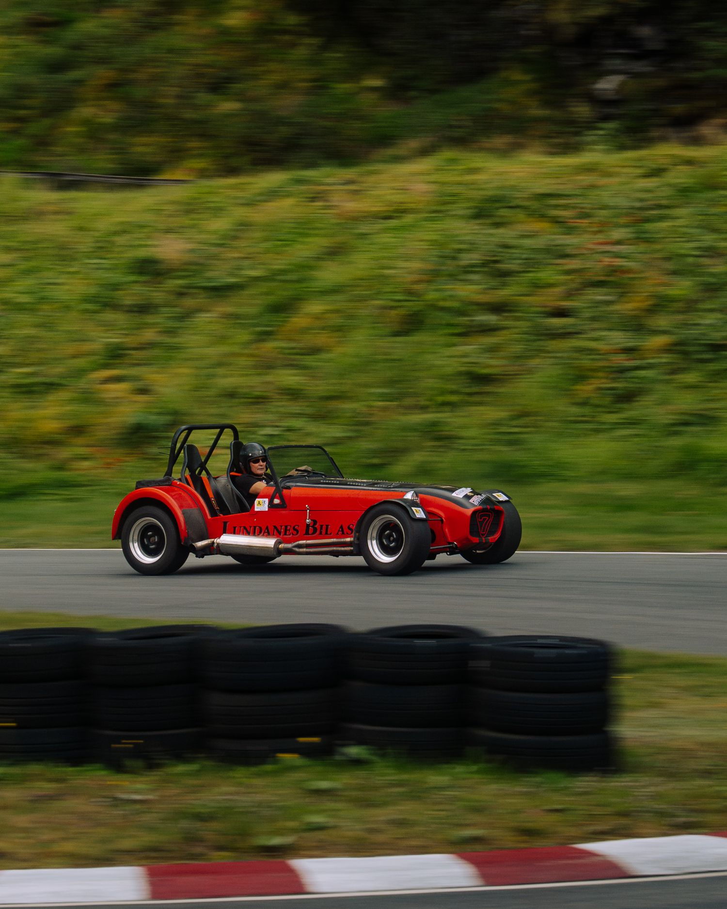 Red vintage-style sports car racing on a track with stacked tires along the curve during a motorsport event.