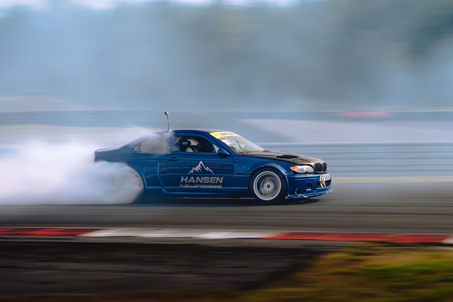 A blue BMW drifts across a racetrack creating smoke trails during a motorsport event.