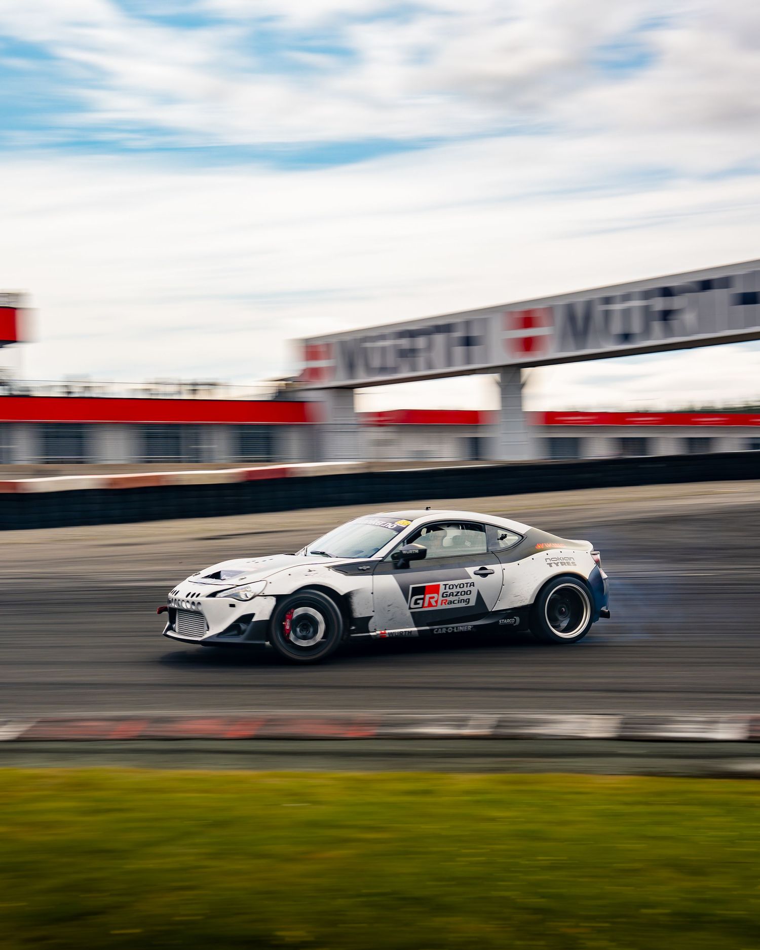 Three motion-blurred shots of racing cars speeding past on a racetrack with grass and cloudy skies in the background.