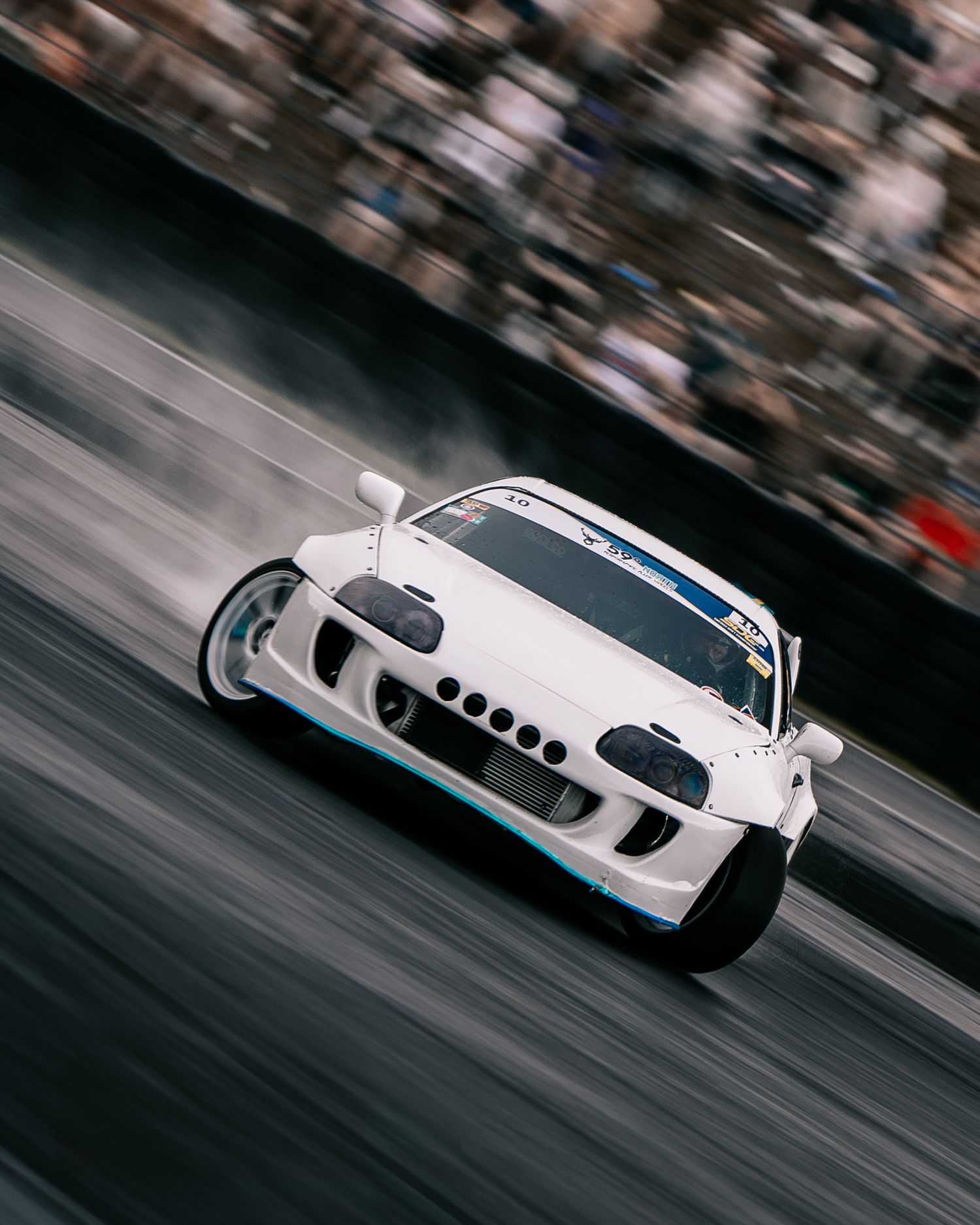 A white sports car racing at high speed on a track with motion blur and spectators in the background.