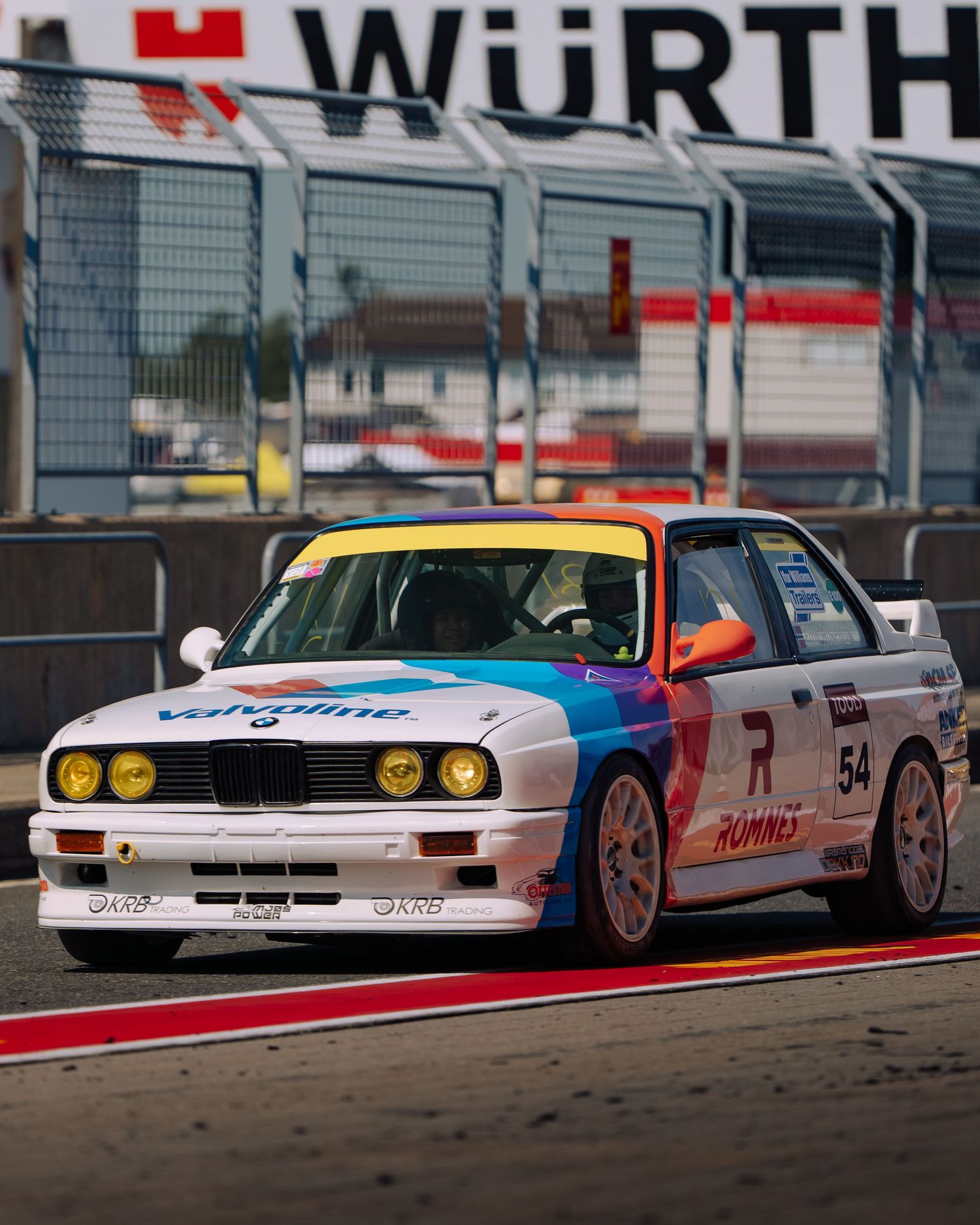 A vintage BMW E30 M3 race car in white, red and blue livery racing on a track with Wurth signage in the background.
