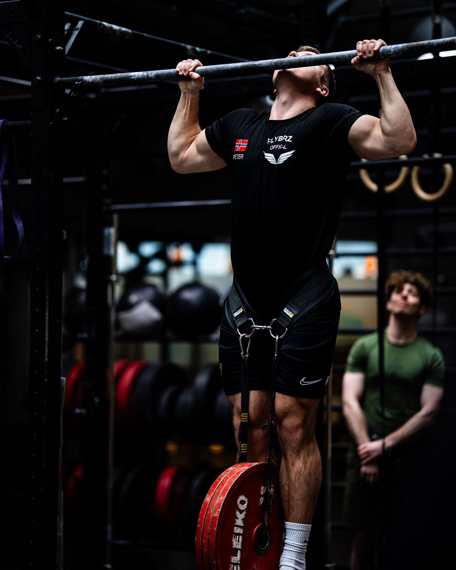 Athlete in black tank top performing pull-up with weight belt in indoor gym setting.