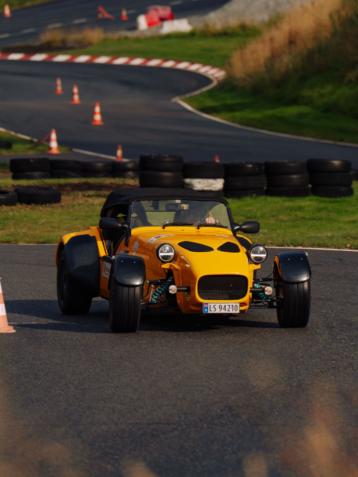 A bright yellow vintage Lotus Super Seven sports car racing around a track marked with orange cones.