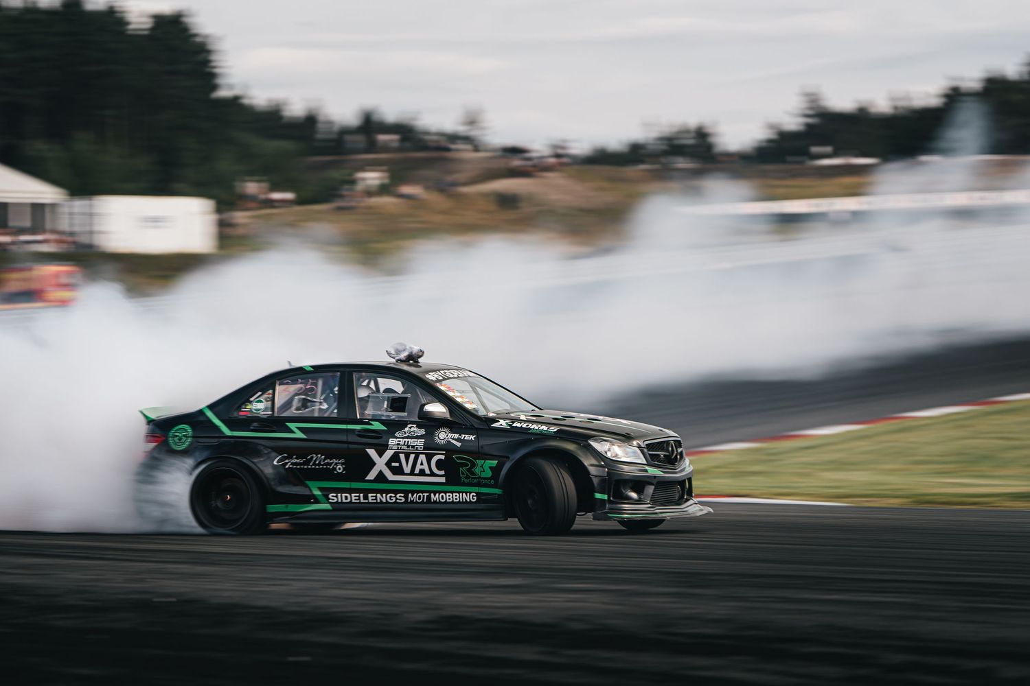 Black BMW M3 drifts around a racetrack creating smoke from burning tires during a motorsport event.
