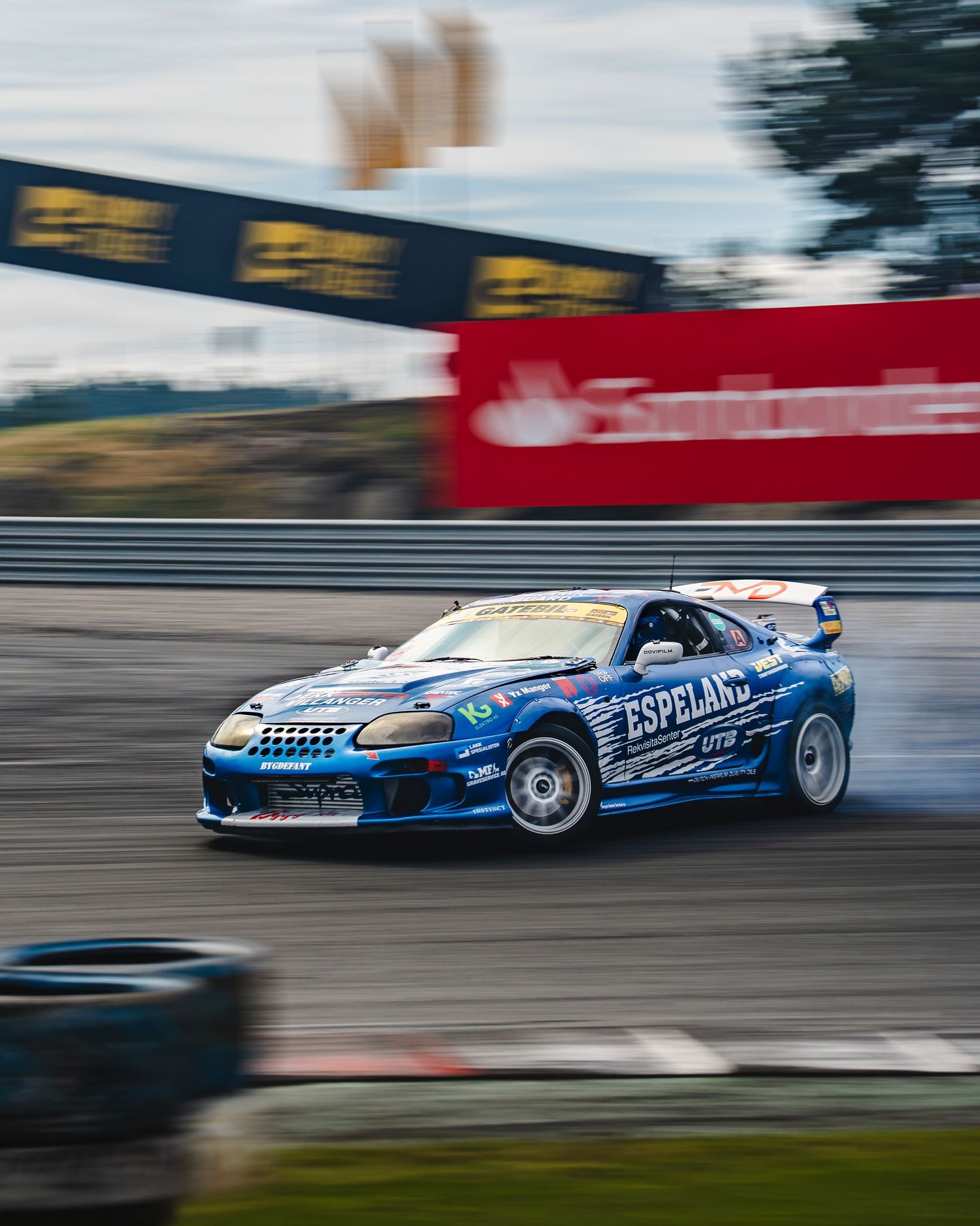 Motion-blurred racing car with blue and white livery drifts around track corner during professional drift competition.