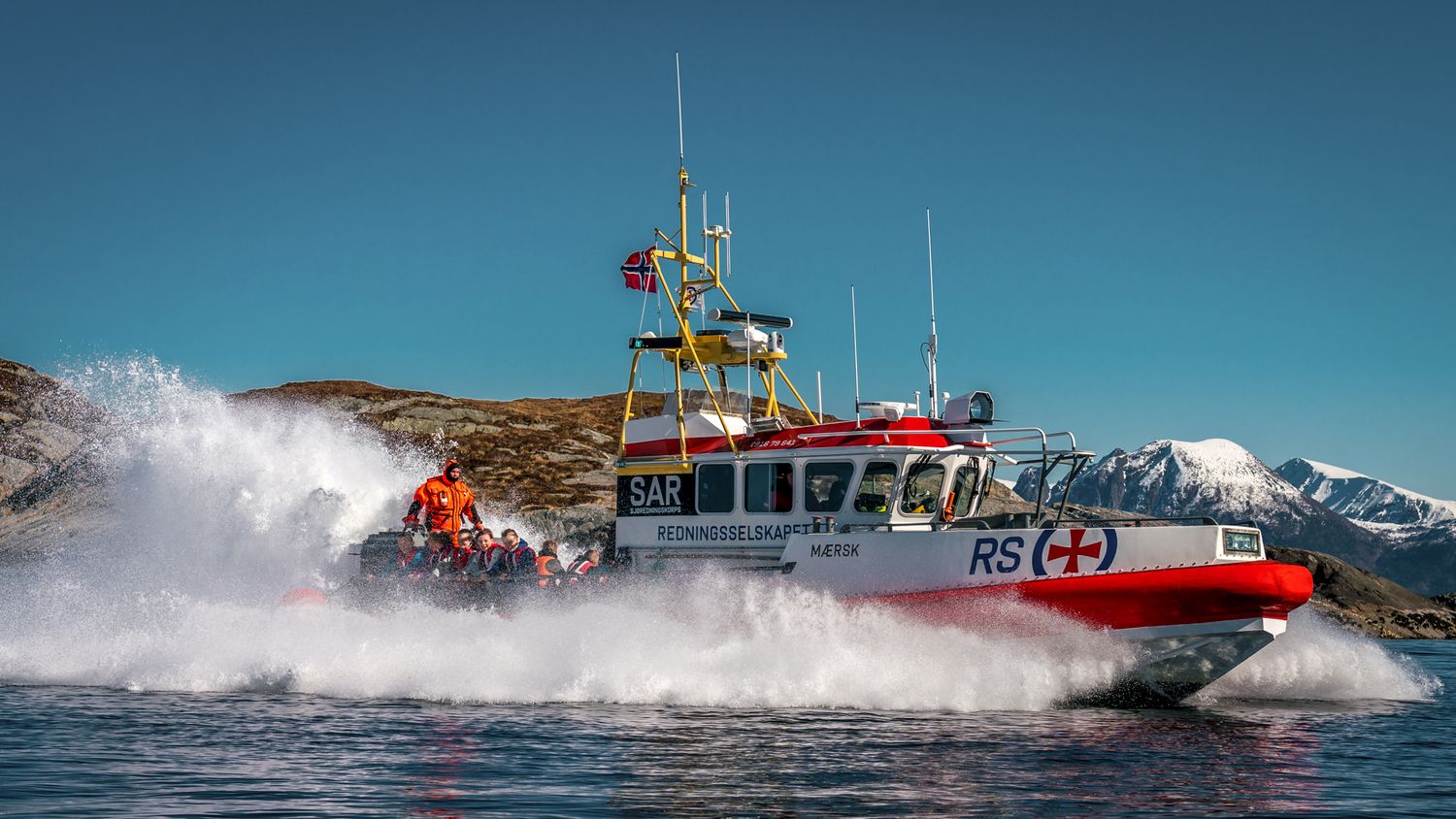 Red rescue vessel creating dramatic spray while navigating through choppy northern waters.