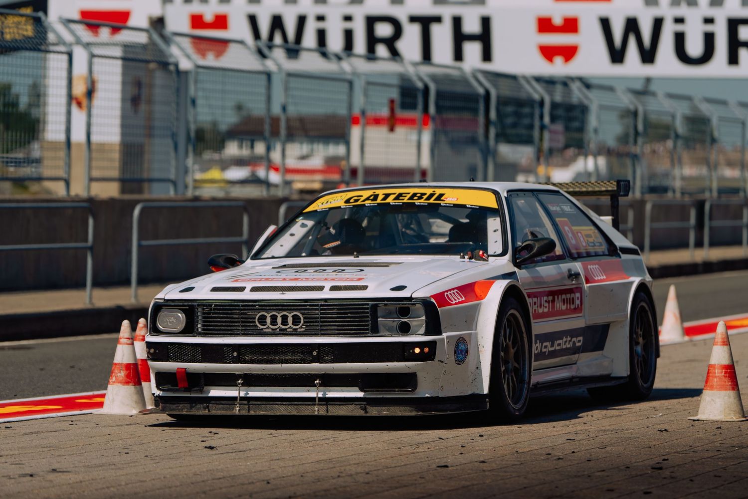 Vintage racing cars including an Audi Quattro and other classic vehicles lined up on a racetrack with safety barriers.