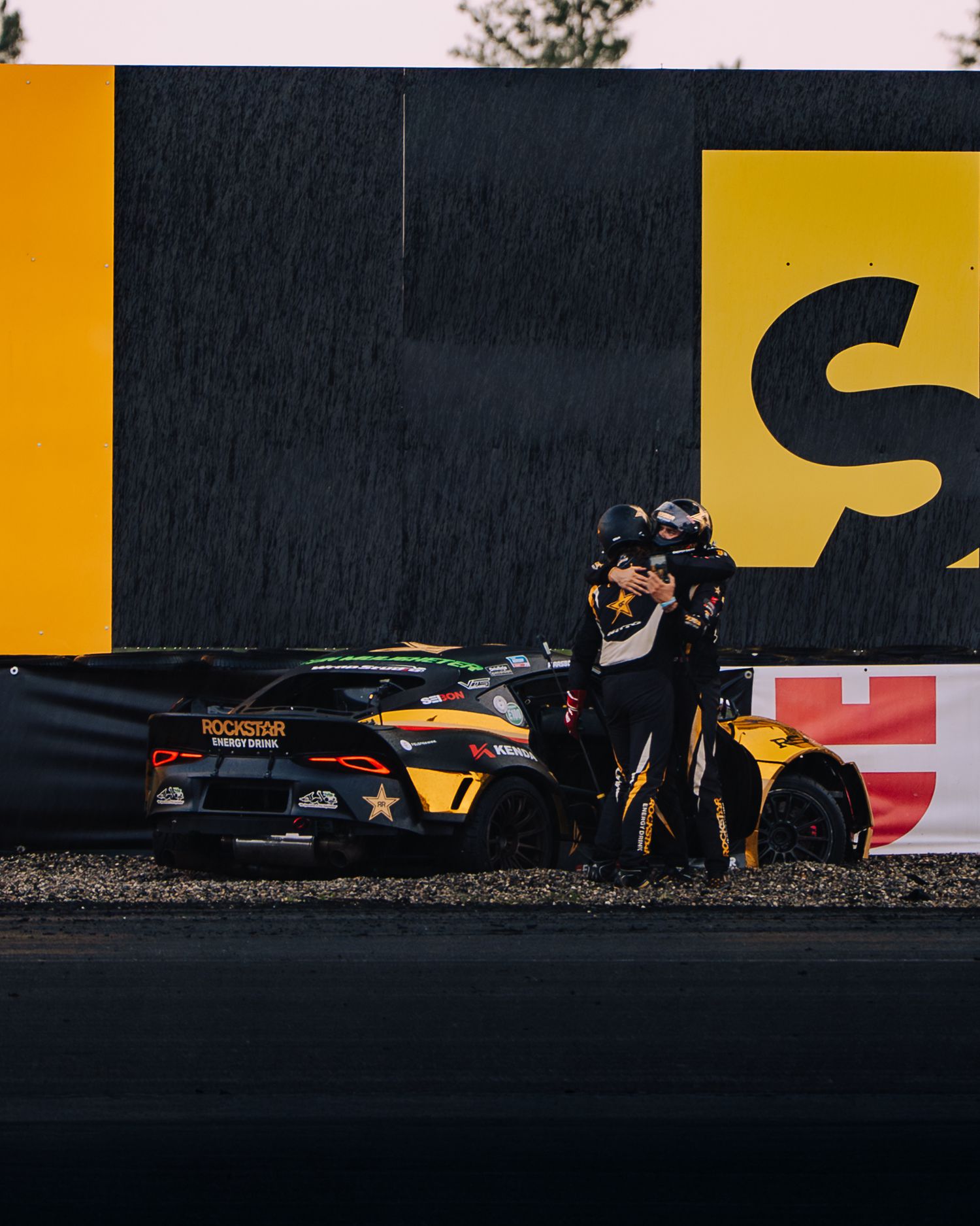 A photographer captures a black sports car against a yellow and black wall in dramatic rainy lighting.
