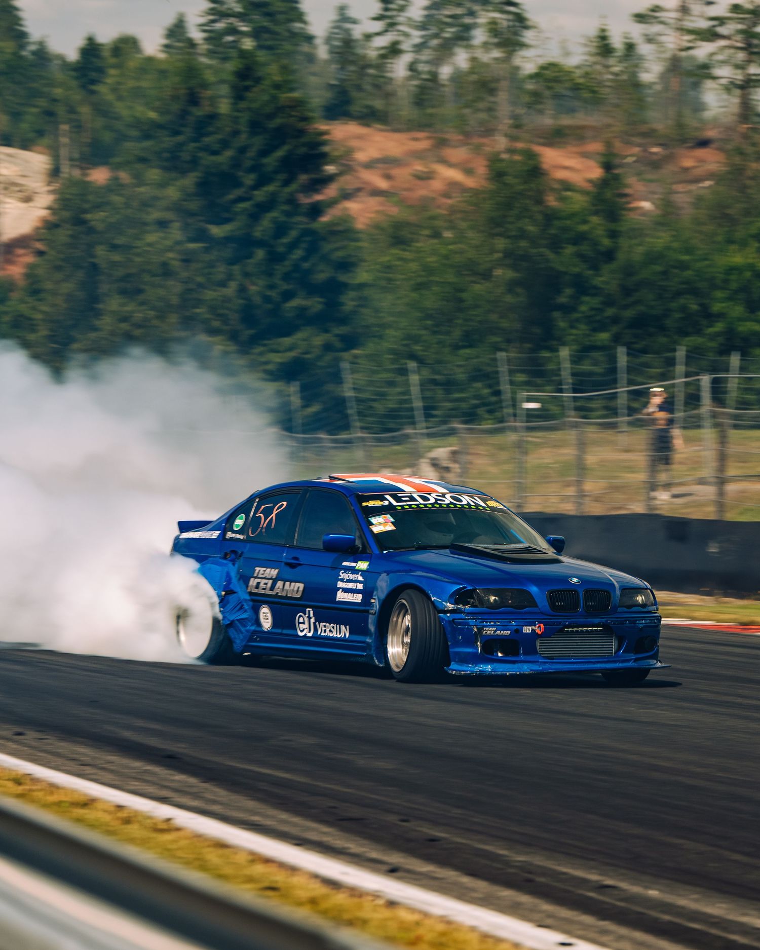 A blue BMW drift car creates smoke while drifting sideways on a racetrack surrounded by evergreen trees.