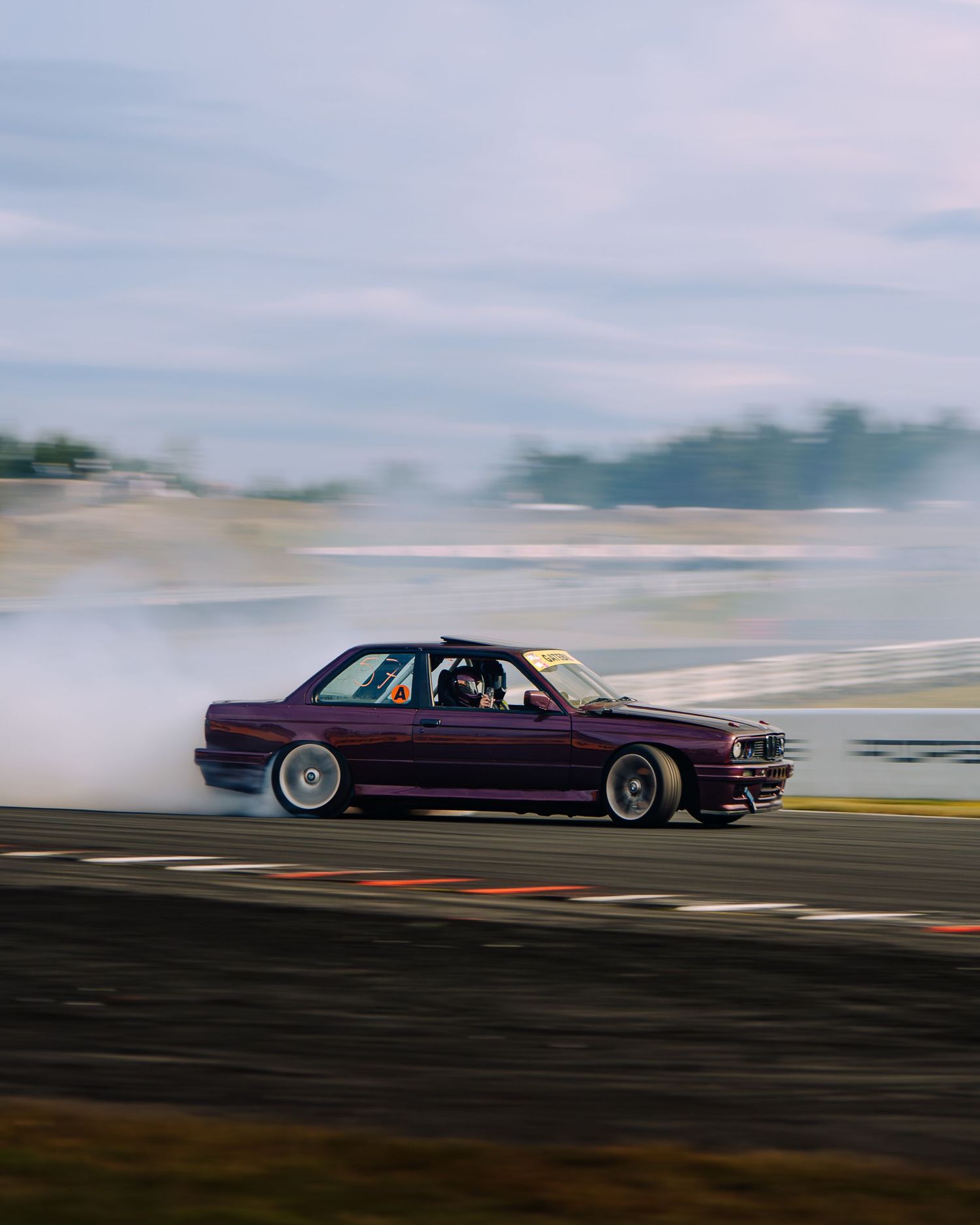 A purple BMW E30 drifting on a race track with tire smoke trailing behind it during a motorsport event.
