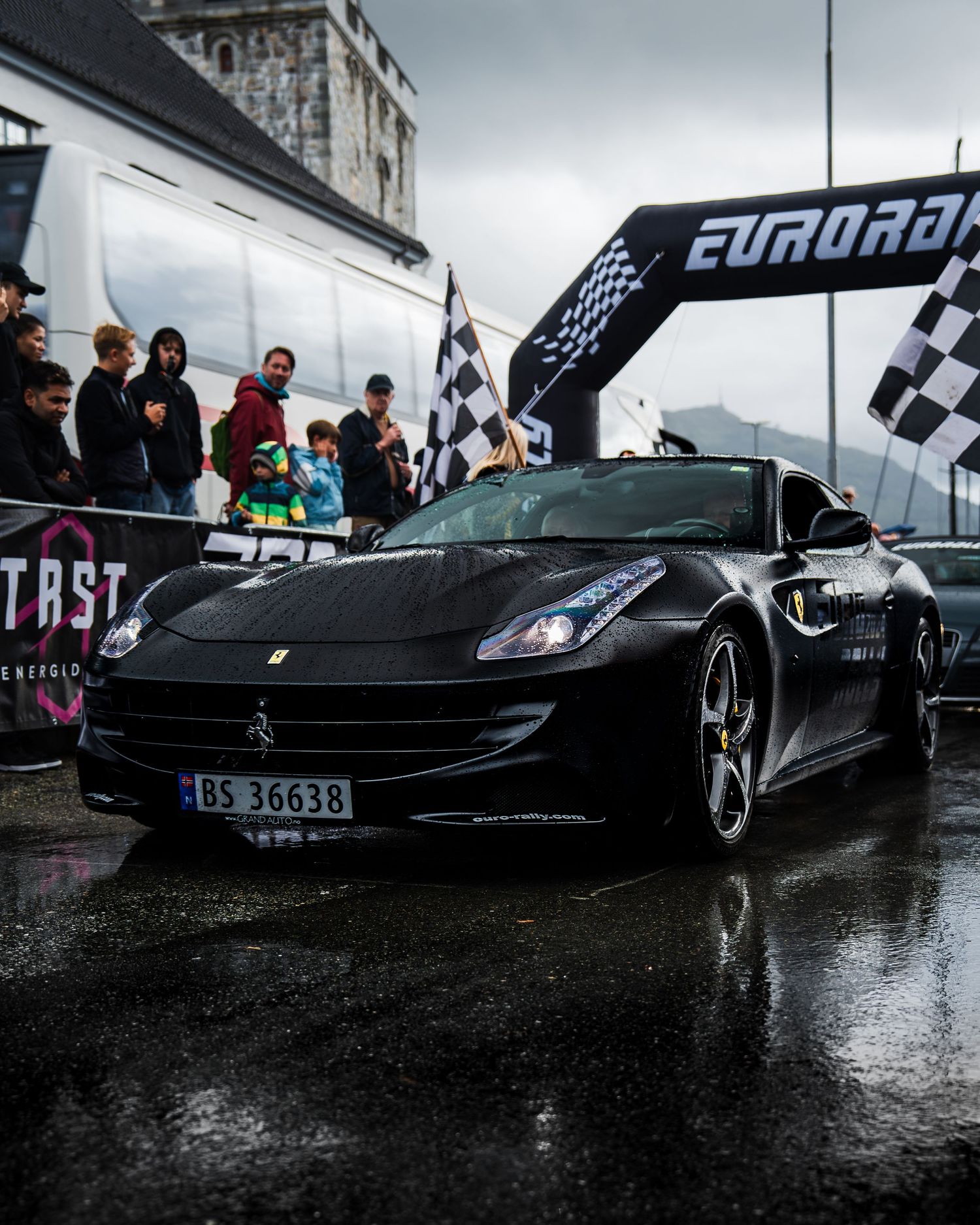 Black Ferrari California passing under Eurorally finish line arch on wet pavement.