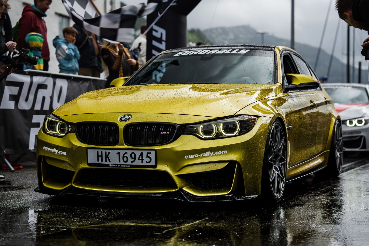 Gold BMW M4 with aggressive styling and LED headlights at rainy Eurorally event.