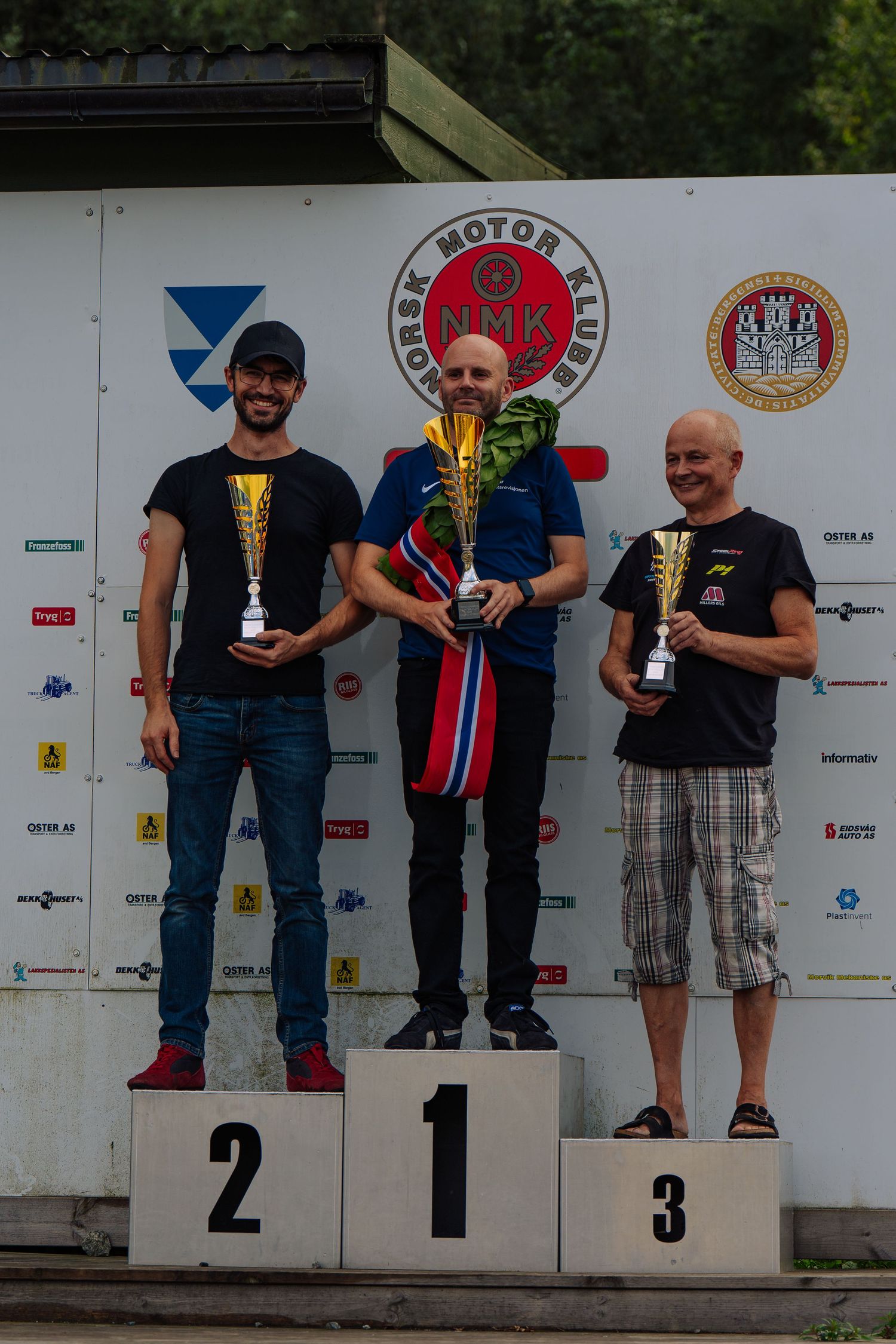 Winners stand on a podium displaying trophies at a motorsport competition award ceremony.
