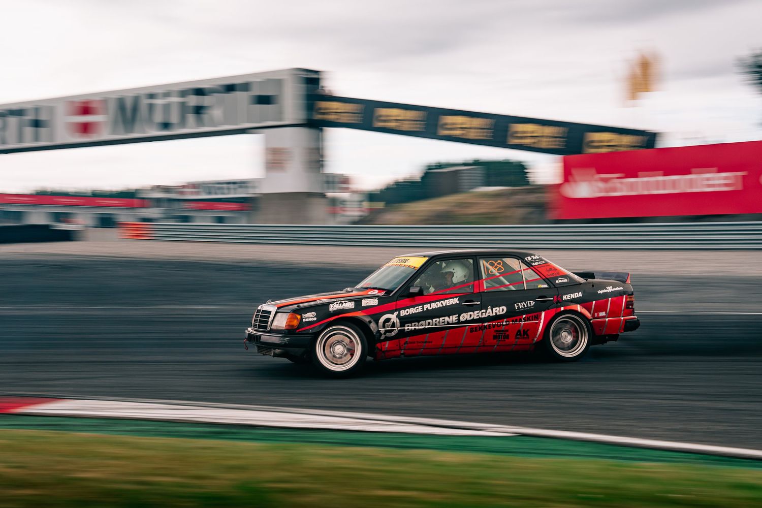 Red race car speeds past on a racetrack with motion blur effect showing dynamic movement and speed.