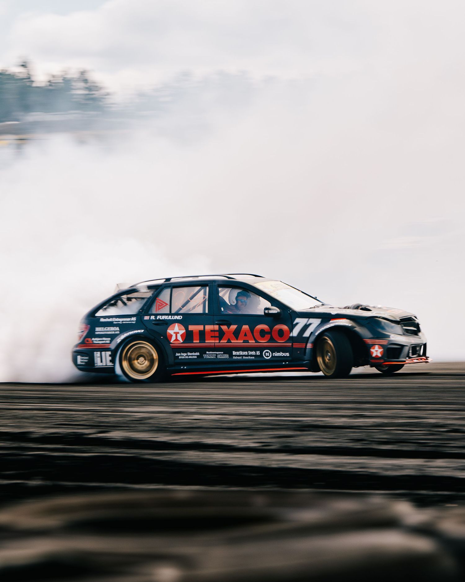 A black Texaco racing car drifts sideways creating smoke while performing a burnout on a track.