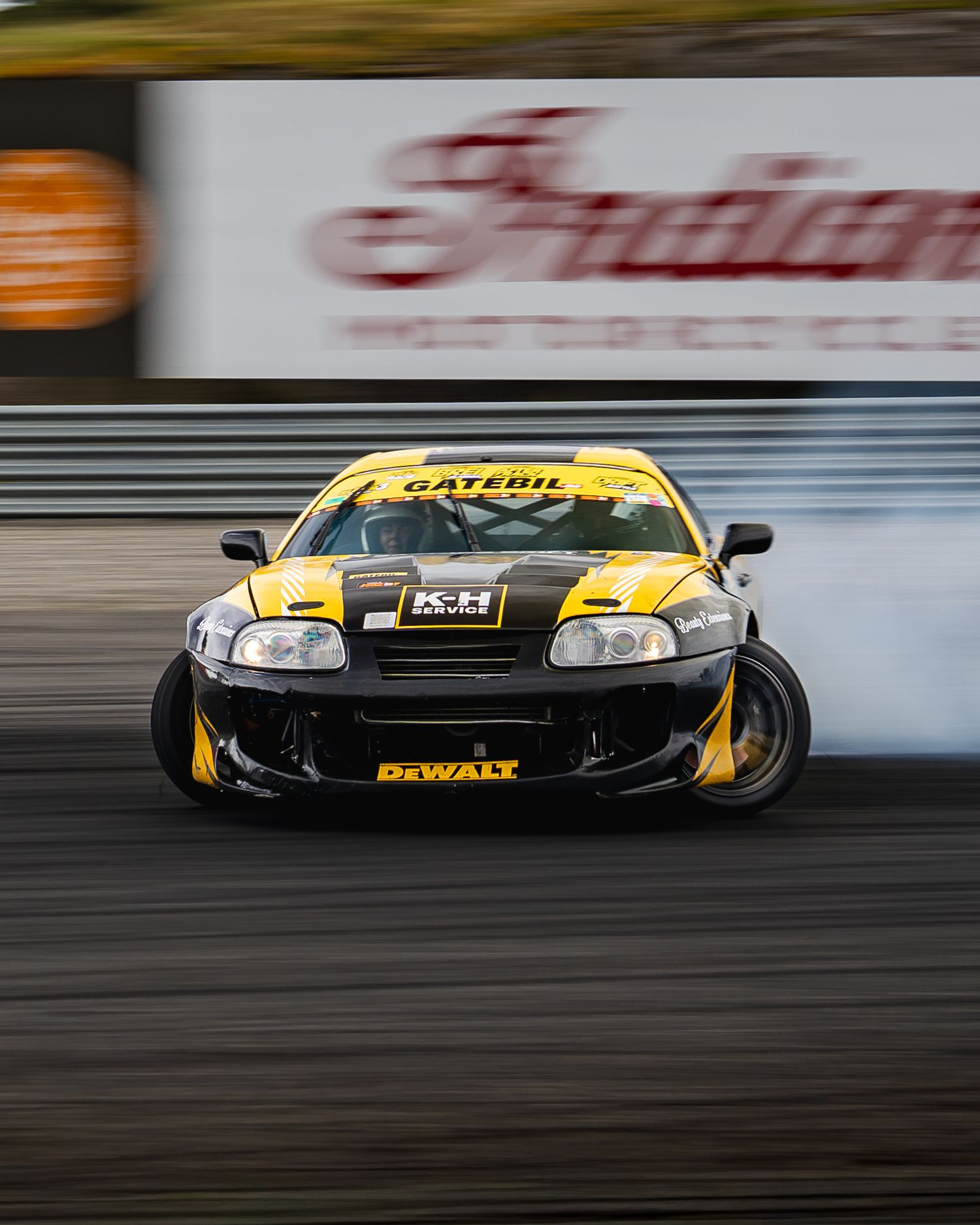 A yellow and black race car drifts sideways on a racetrack with motion blur in the background.
