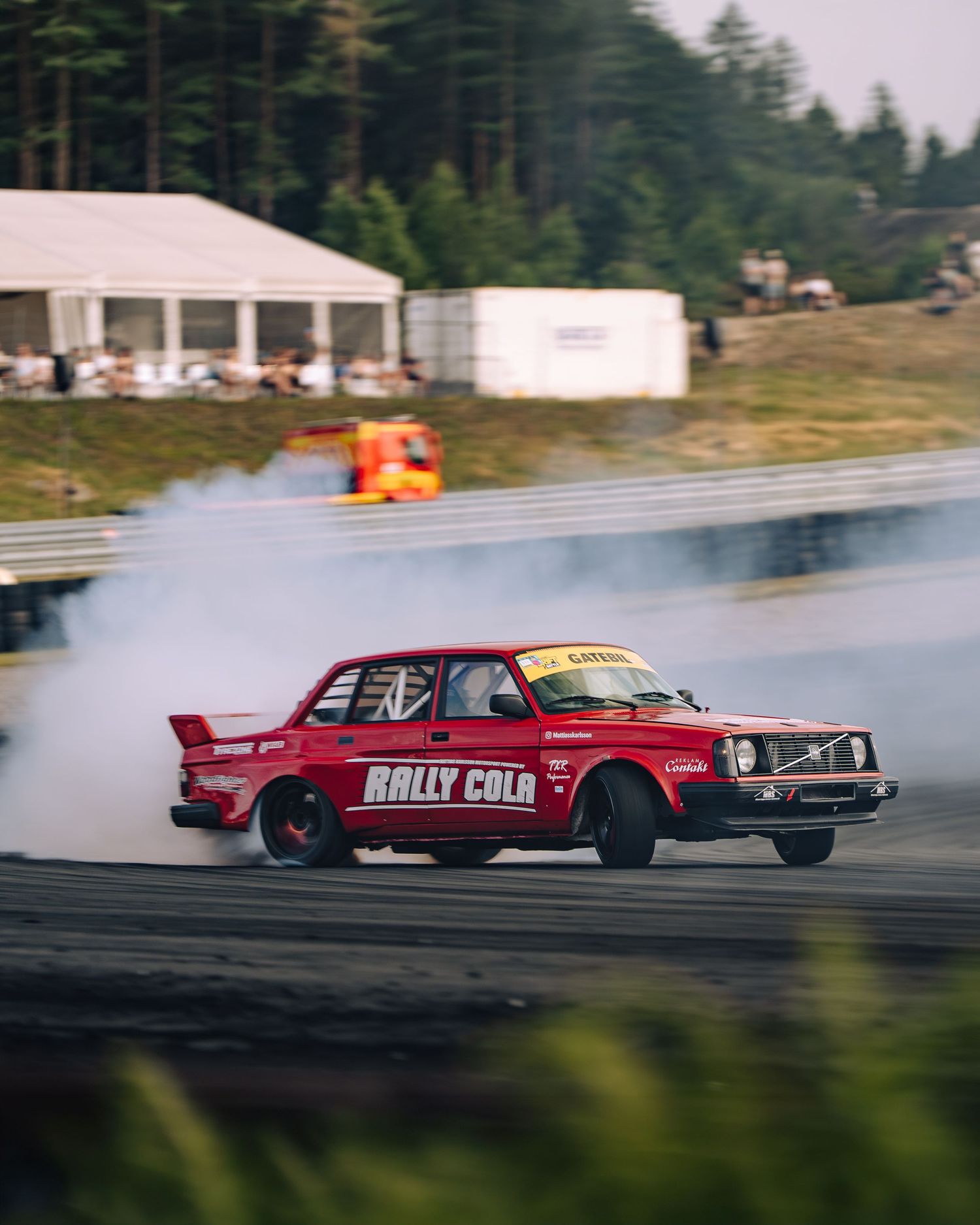 A red vintage Volvo performs a drift with smoke billowing from its tires on a racetrack during a rally event.