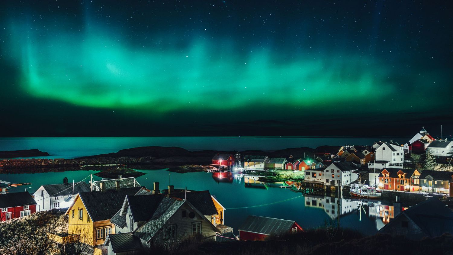 Vibrant aurora borealis glows over a seaside village with colored houses reflecting in the harbor.