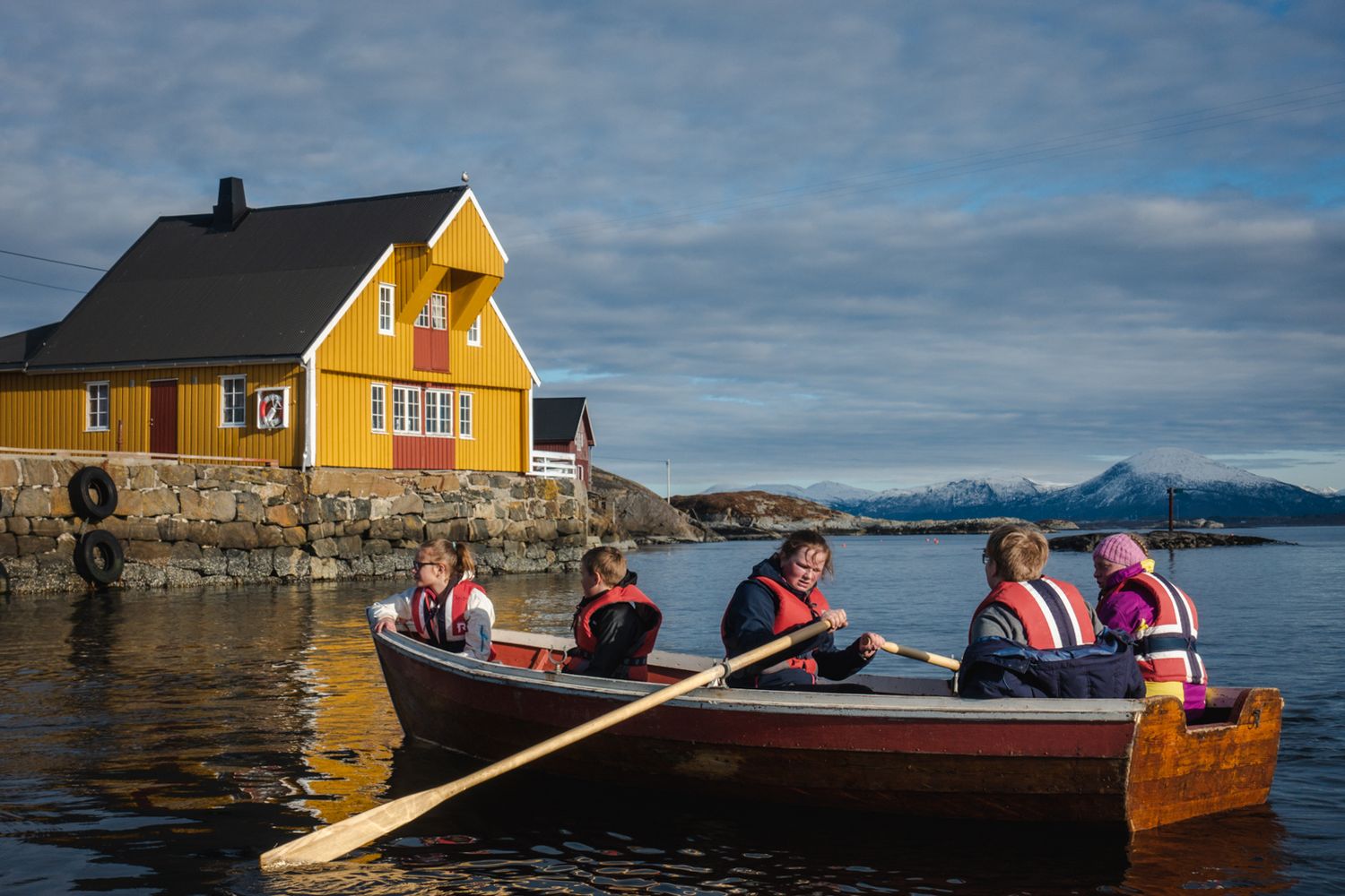 Yellow house overlooks wooden boat with passengers rowing in Nordic coastal waters.