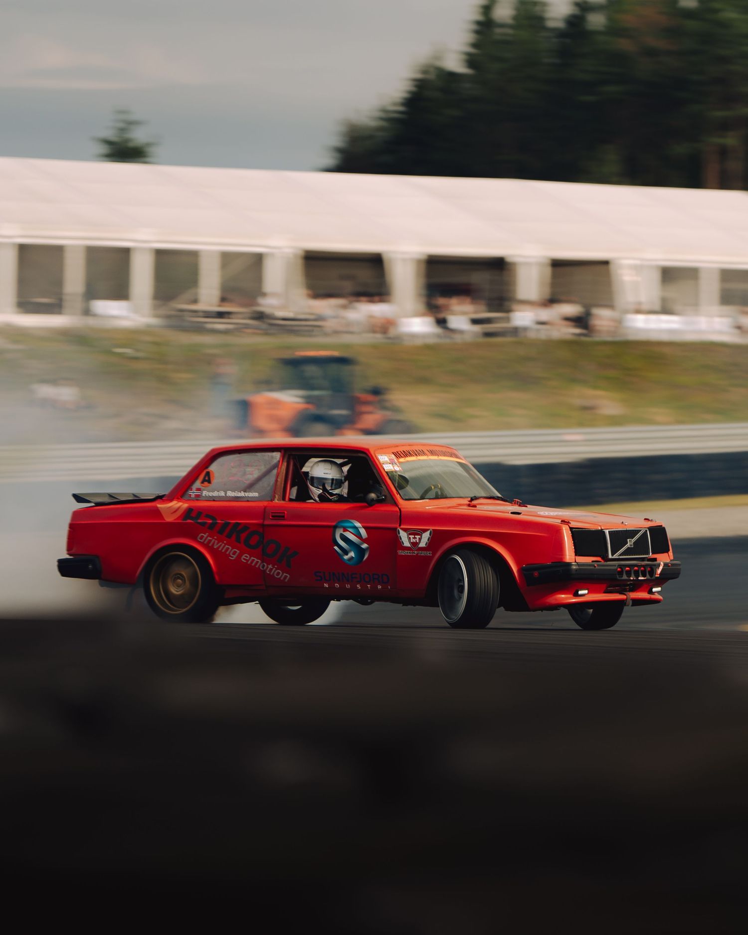 A red Volvo race car drifts sideways on a racetrack with motion blur and white buildings in the background.