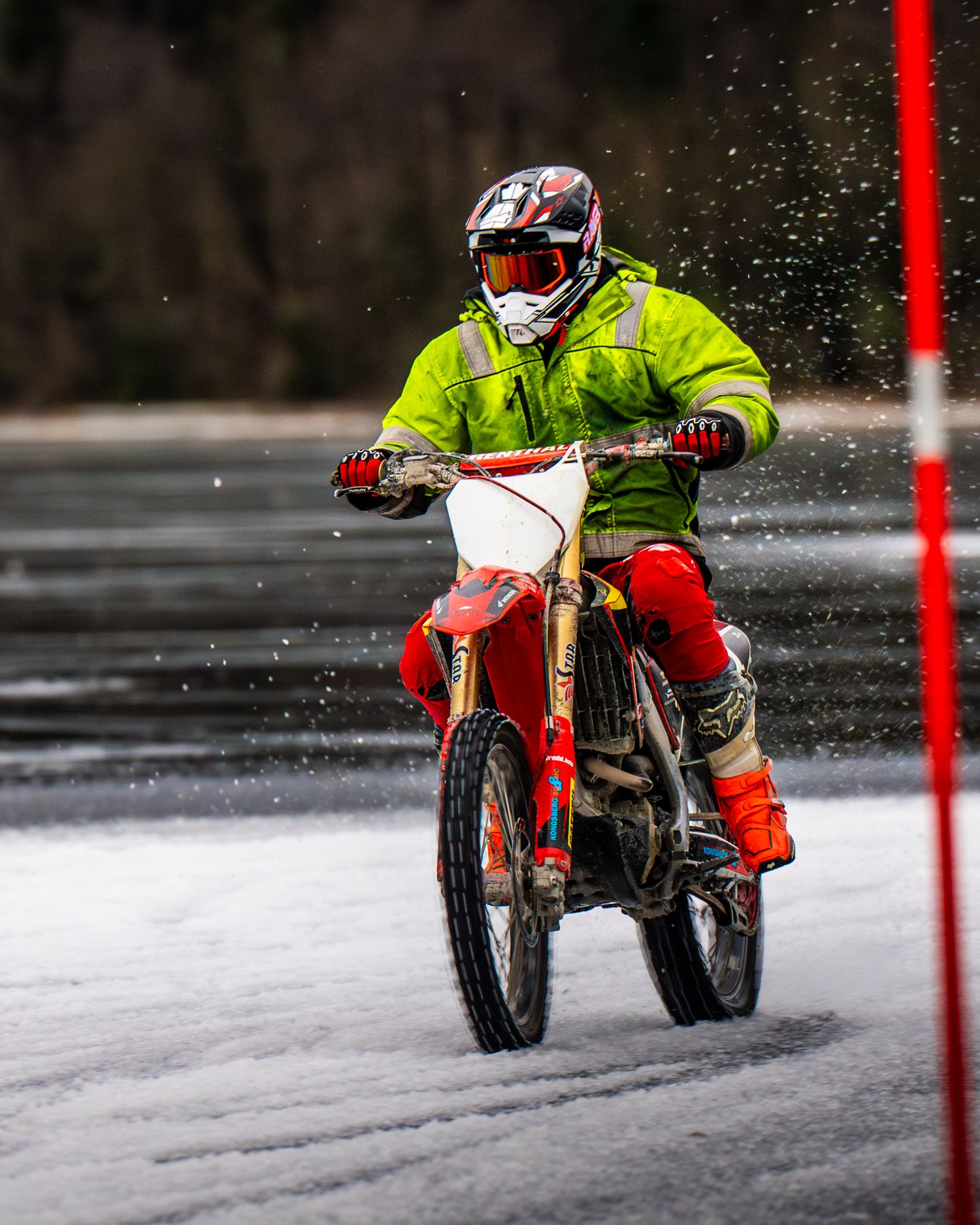 Motorcyclist riding a dirt bike on snowy terrain creates dramatic spray while racing past red markers.
