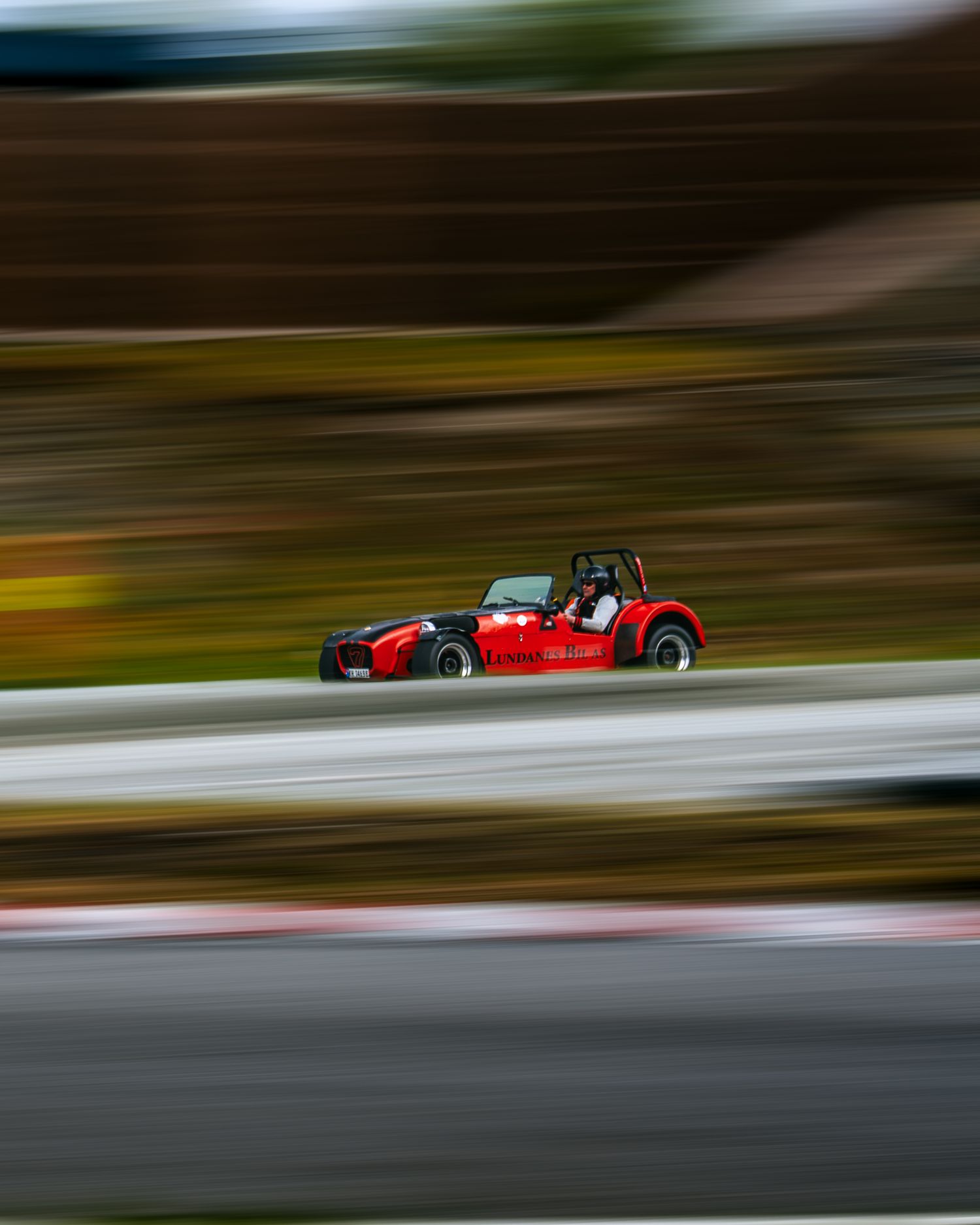 Motion-blurred racing cars speed past on a racetrack with autumn scenery in the background.