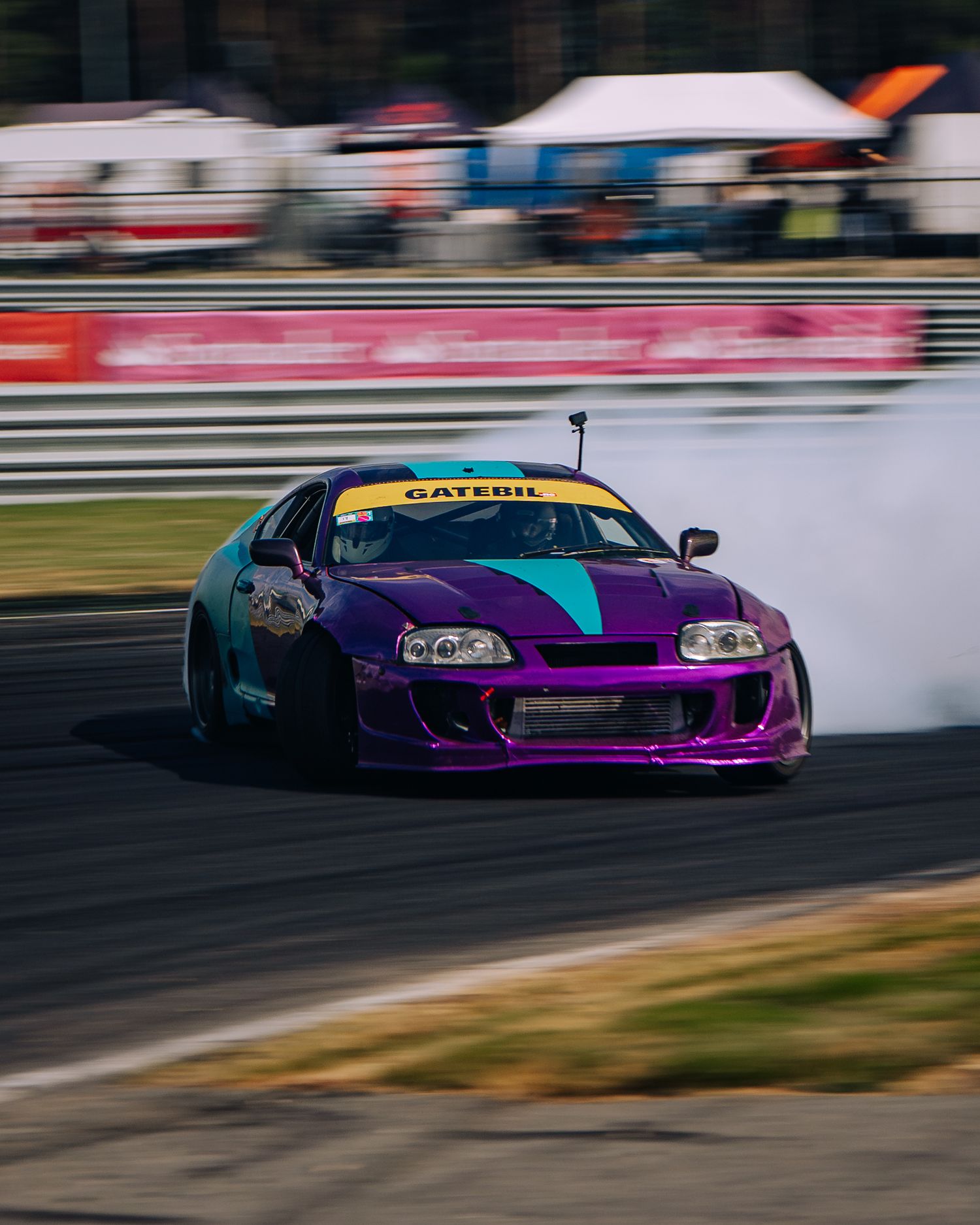 A purple Toyota Supra drifts on a race track with smoke trailing from its tires during a motorsport event.