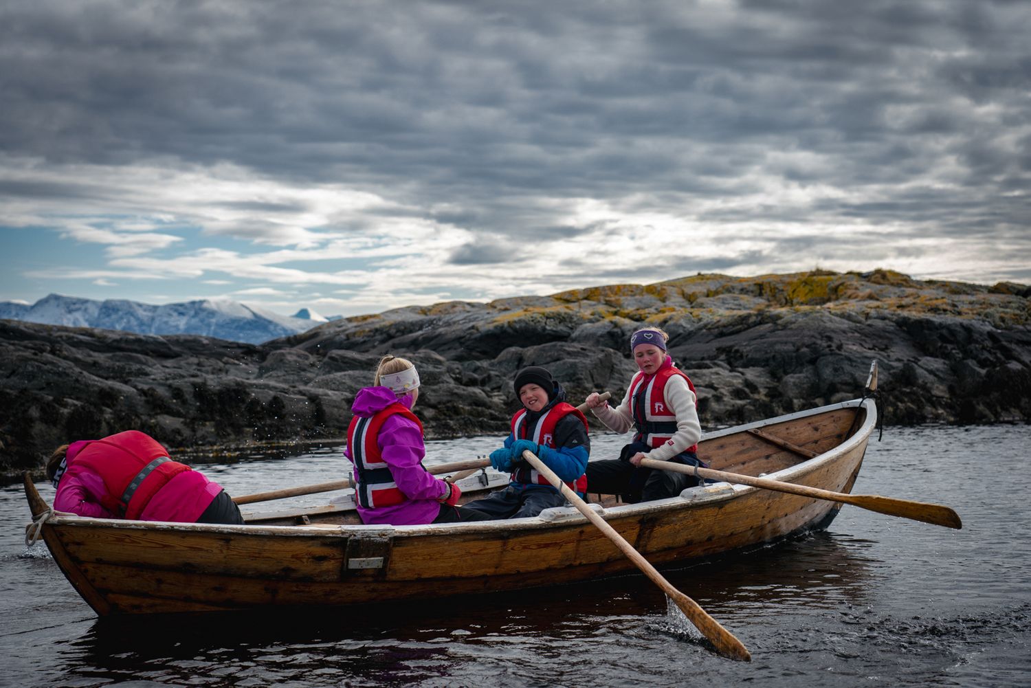 Wooden rowboat with passengers navigating rocky coastal waters under dramatic cloudy skies.