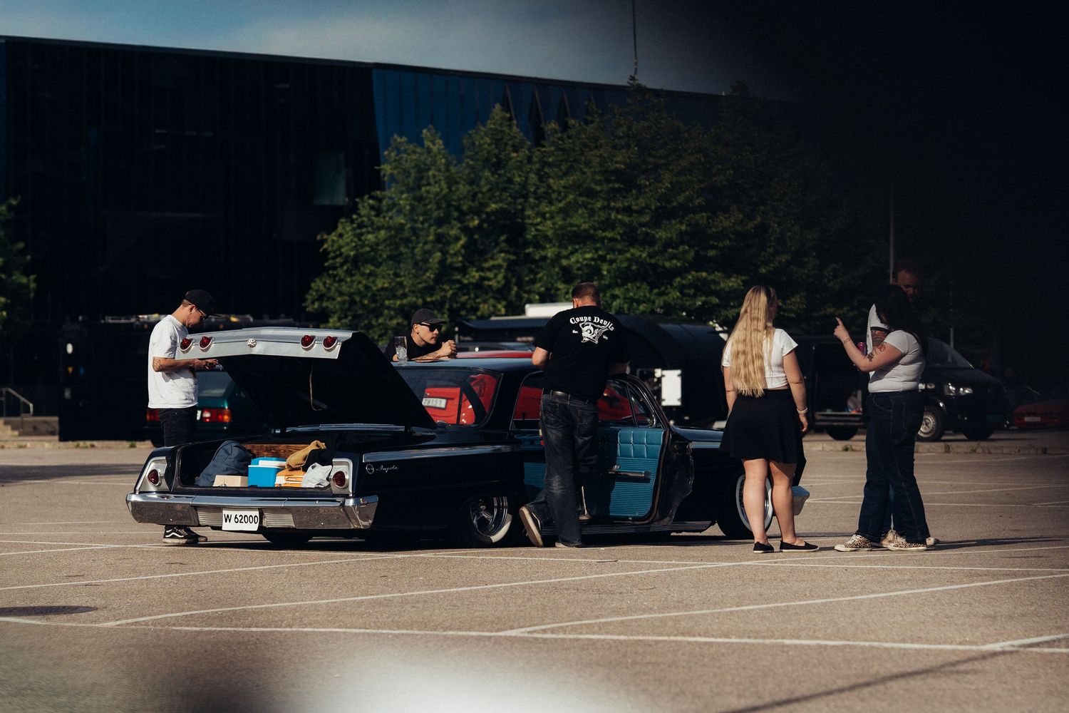 A group of people gathered around an open car trunk in a parking lot during daytime.